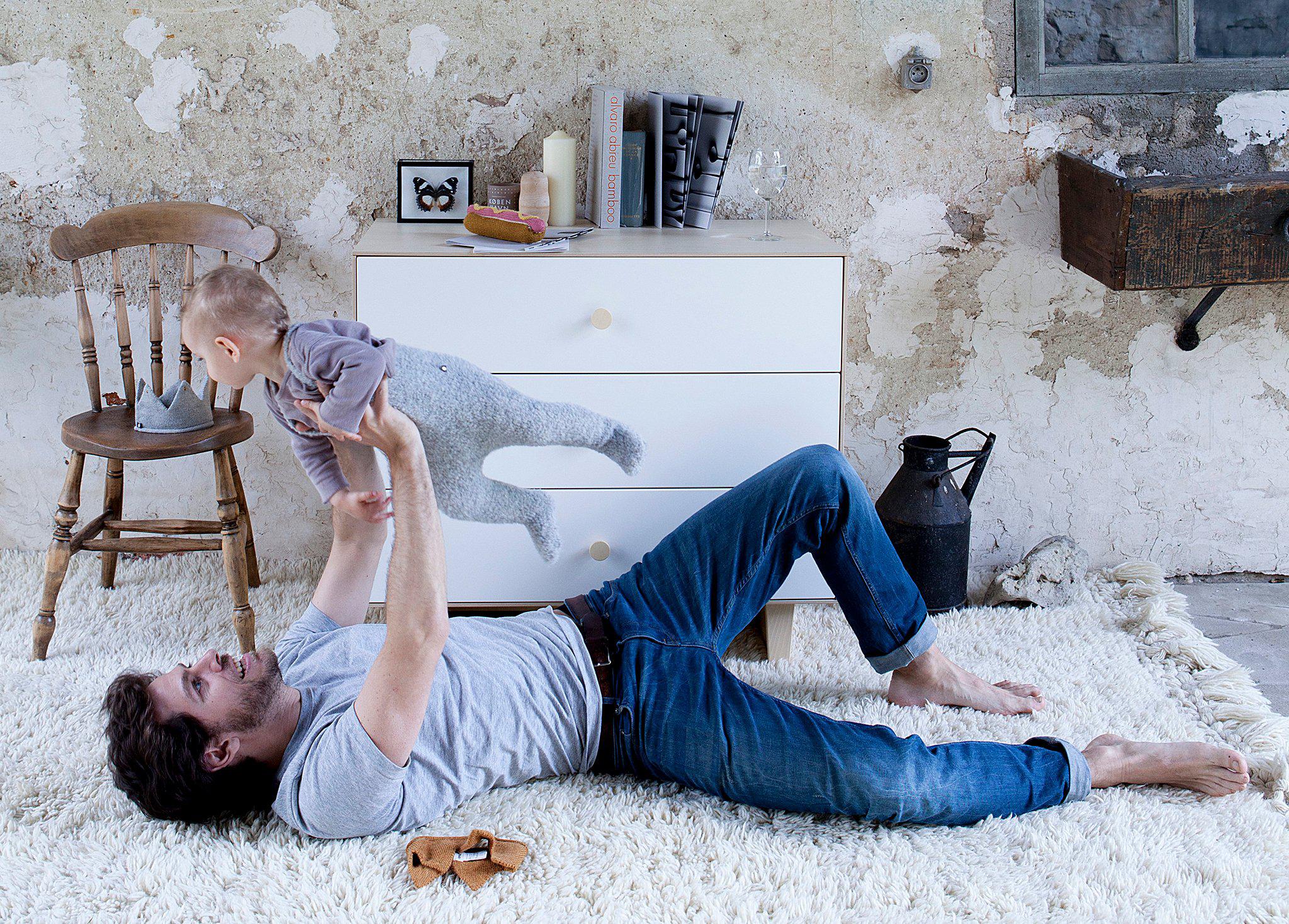 A man lies on a rug, lifting a baby above him in a cozy, rustic room with light-colored walls and furniture.