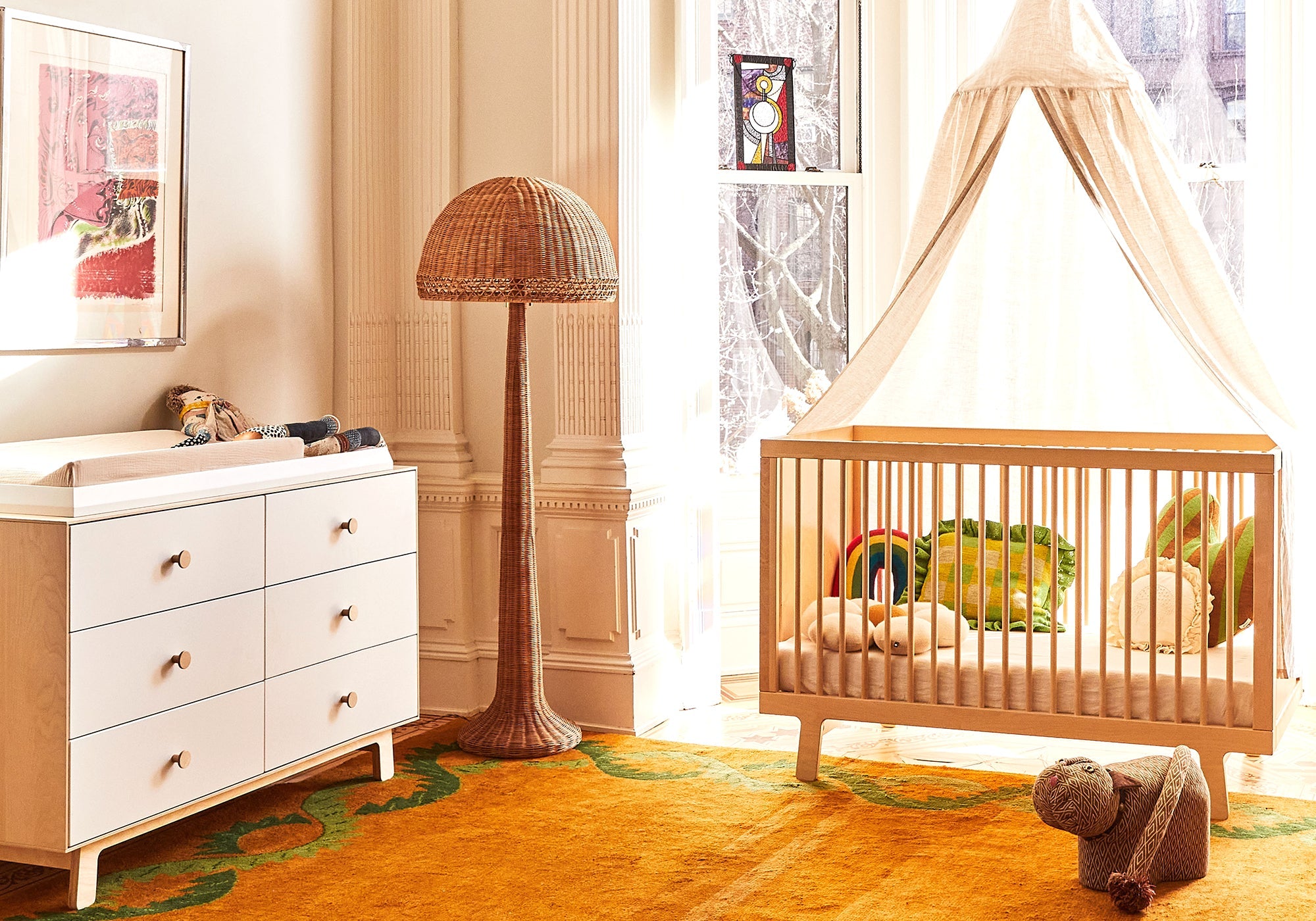 Bright nursery room with a crib, canopy, dresser, rattan lamp, stuffed toys, and a yellow carpet.