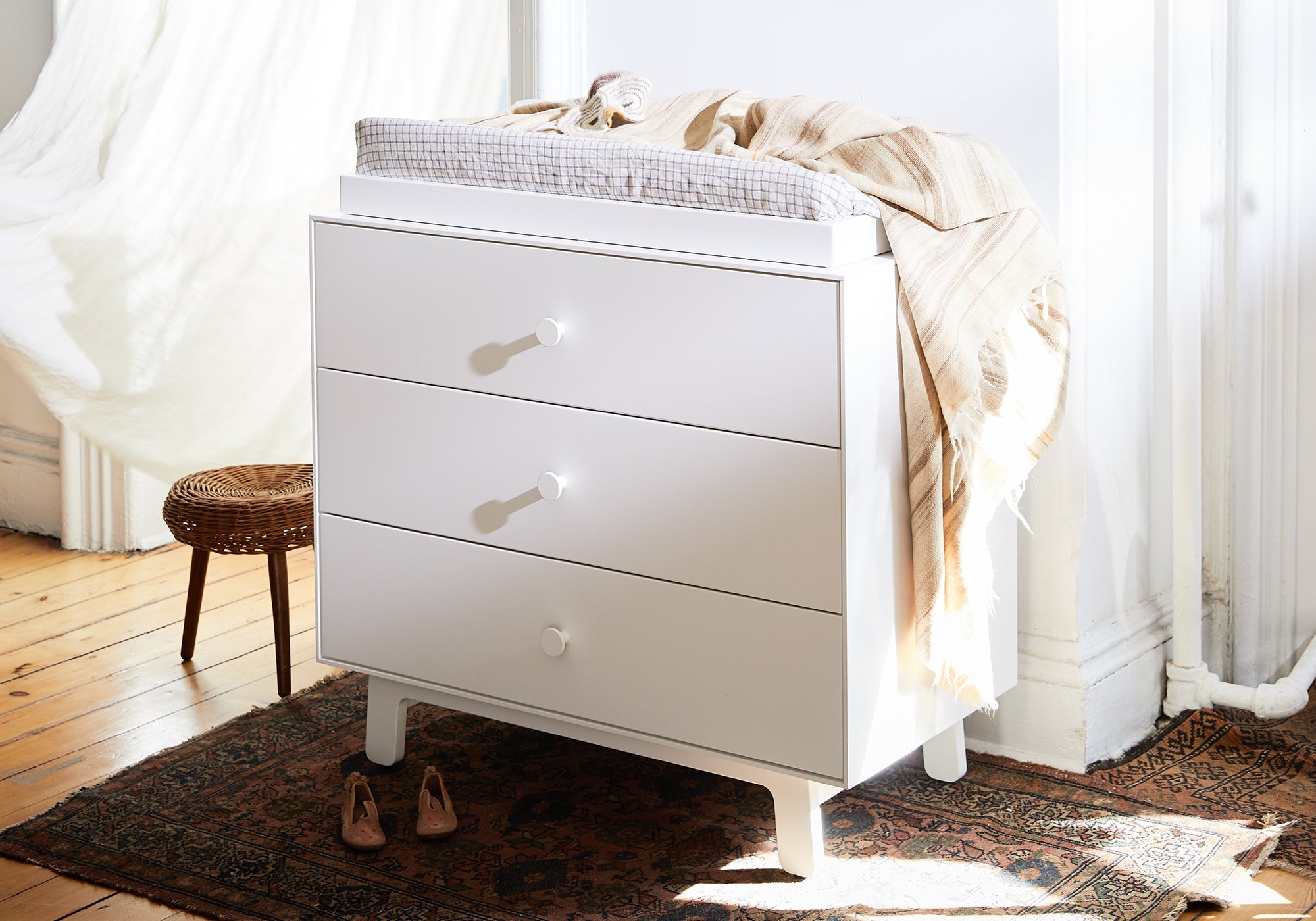 White dresser with three drawers, topped with a checkered changing pad and beige blanket, in a sunlit room.