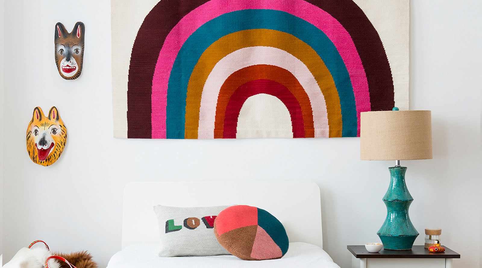 A colorful rainbow tapestry hangs above a bed with playful pillows and a turquoise lamp on a nightstand.