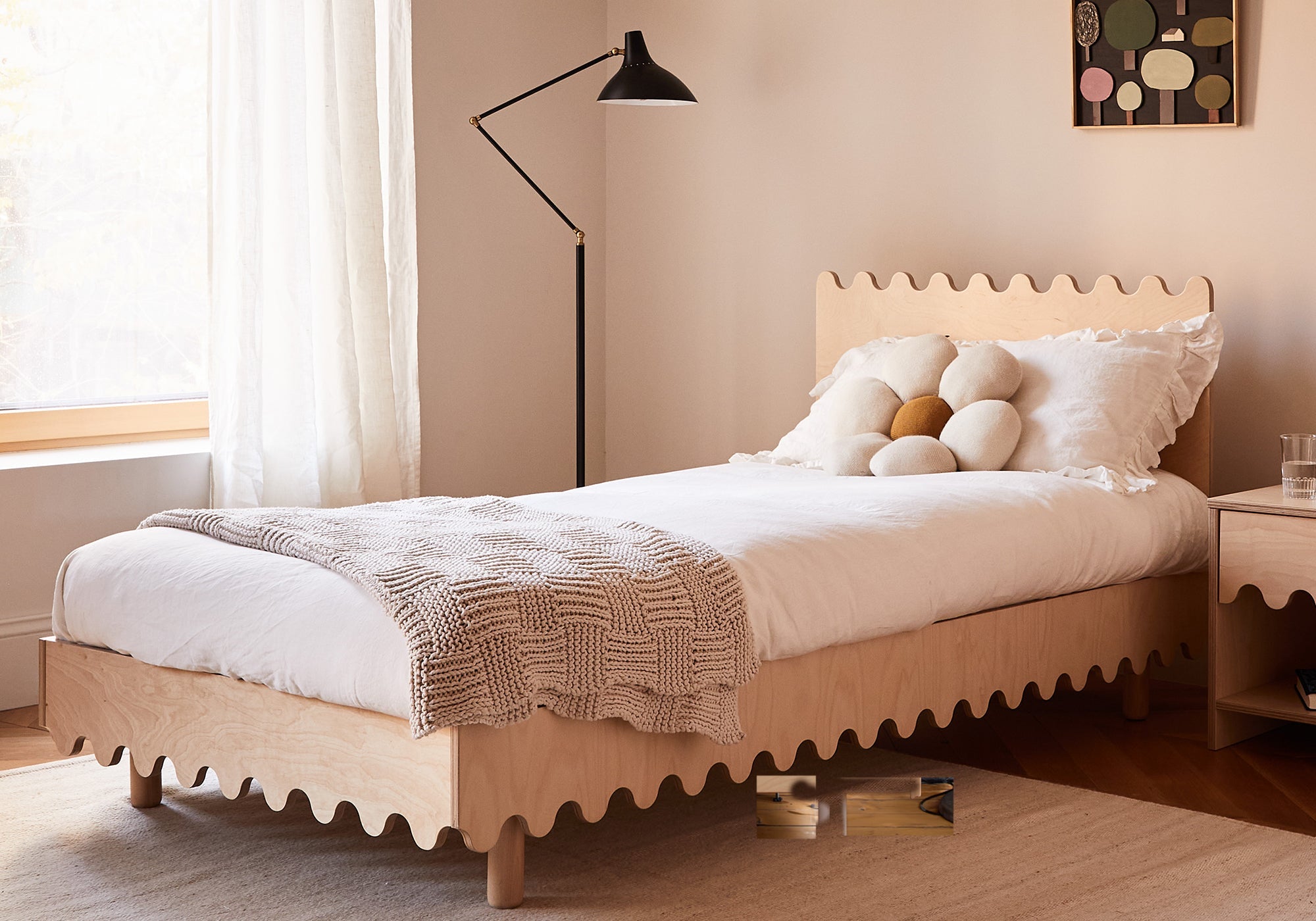 A light wood twin bed with wavy edges, white bedding, and round pillows, beside a floor lamp and nightstand.