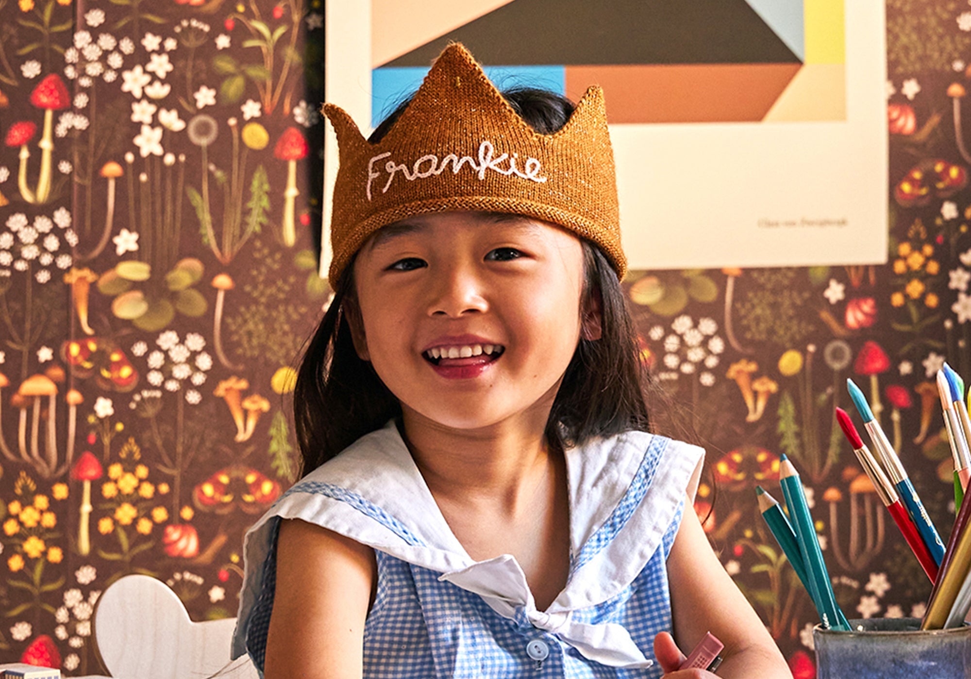 Smiling child in a crown labeled "Frankie," sitting at a desk with pens, against a floral wallpaper background.