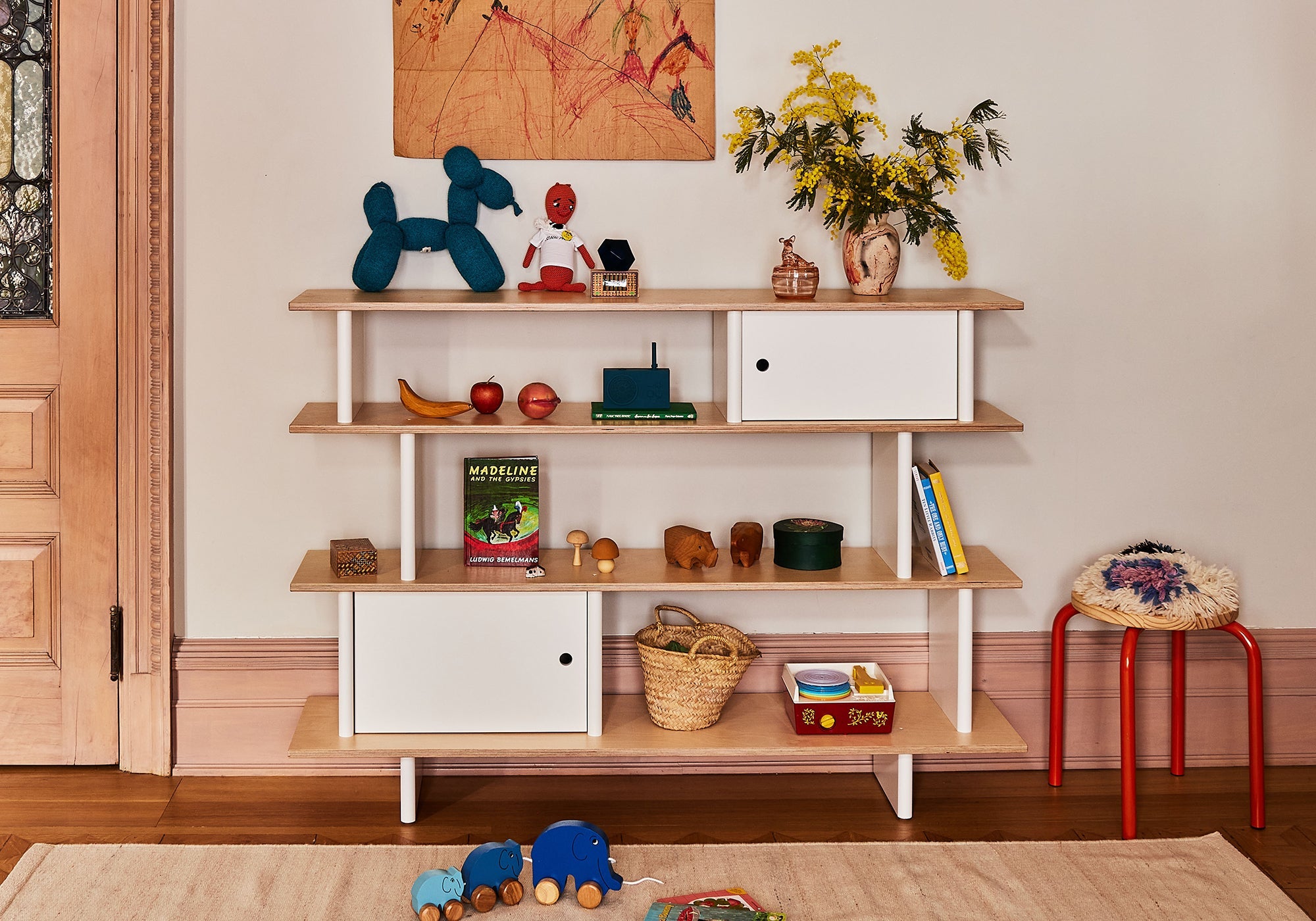 A wooden shelf holds toys, books, plants, and decor, with more toys and a rug on the floor in front.