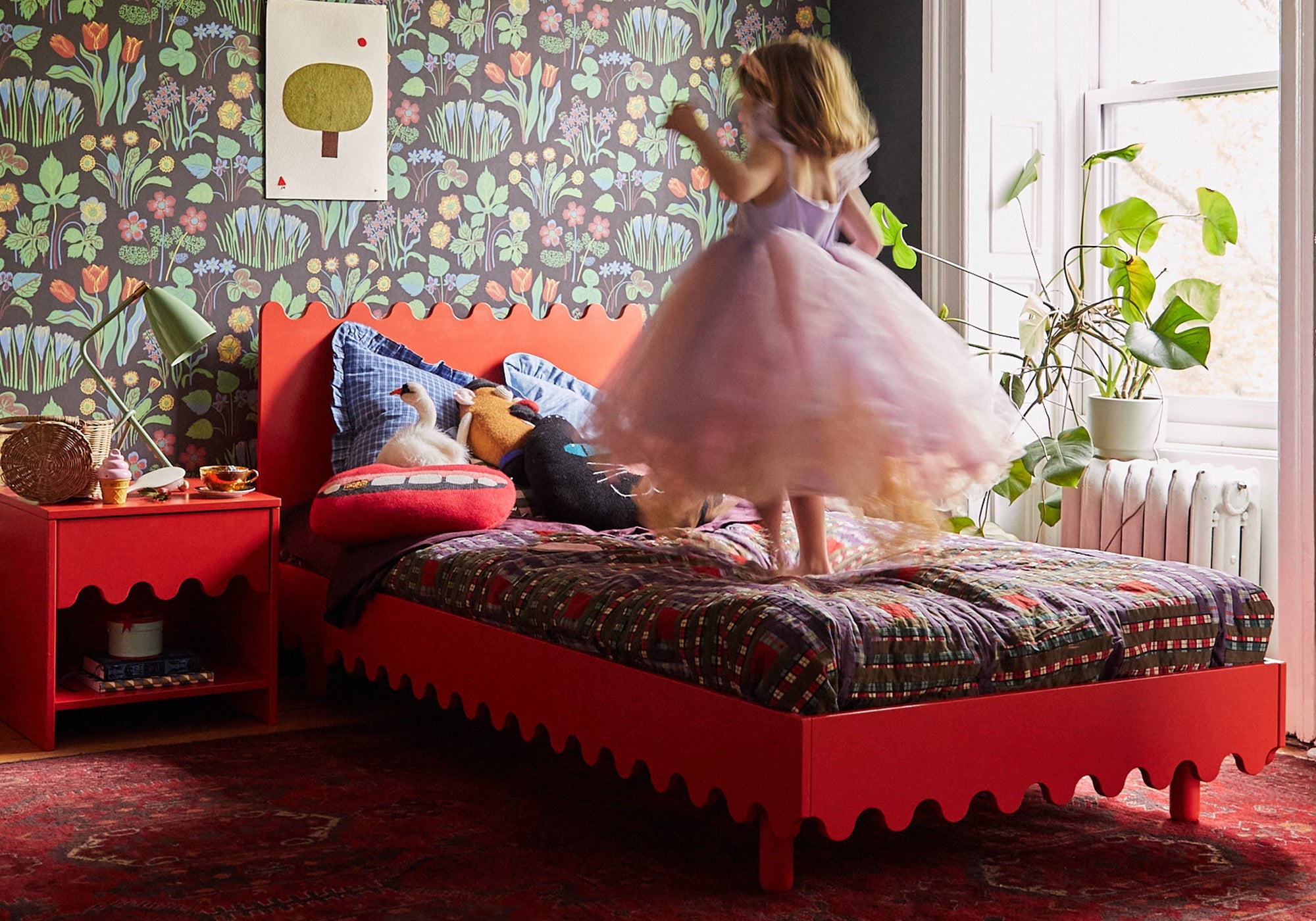 A young girl in a dress jumps on a colorful bed in a vibrant, playful bedroom with floral wallpaper.