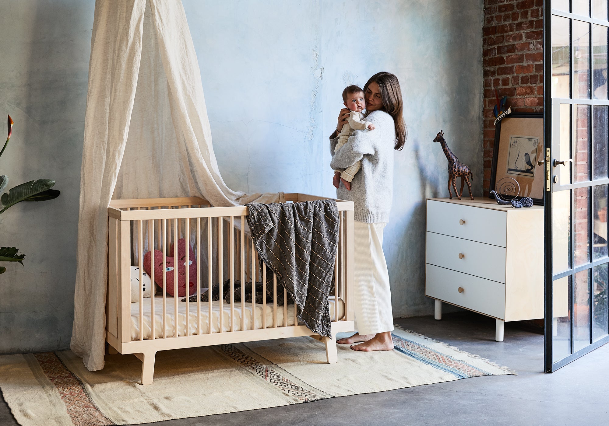 A woman stands by a crib in a cozy, softly lit nursery, holding a baby in her arms.