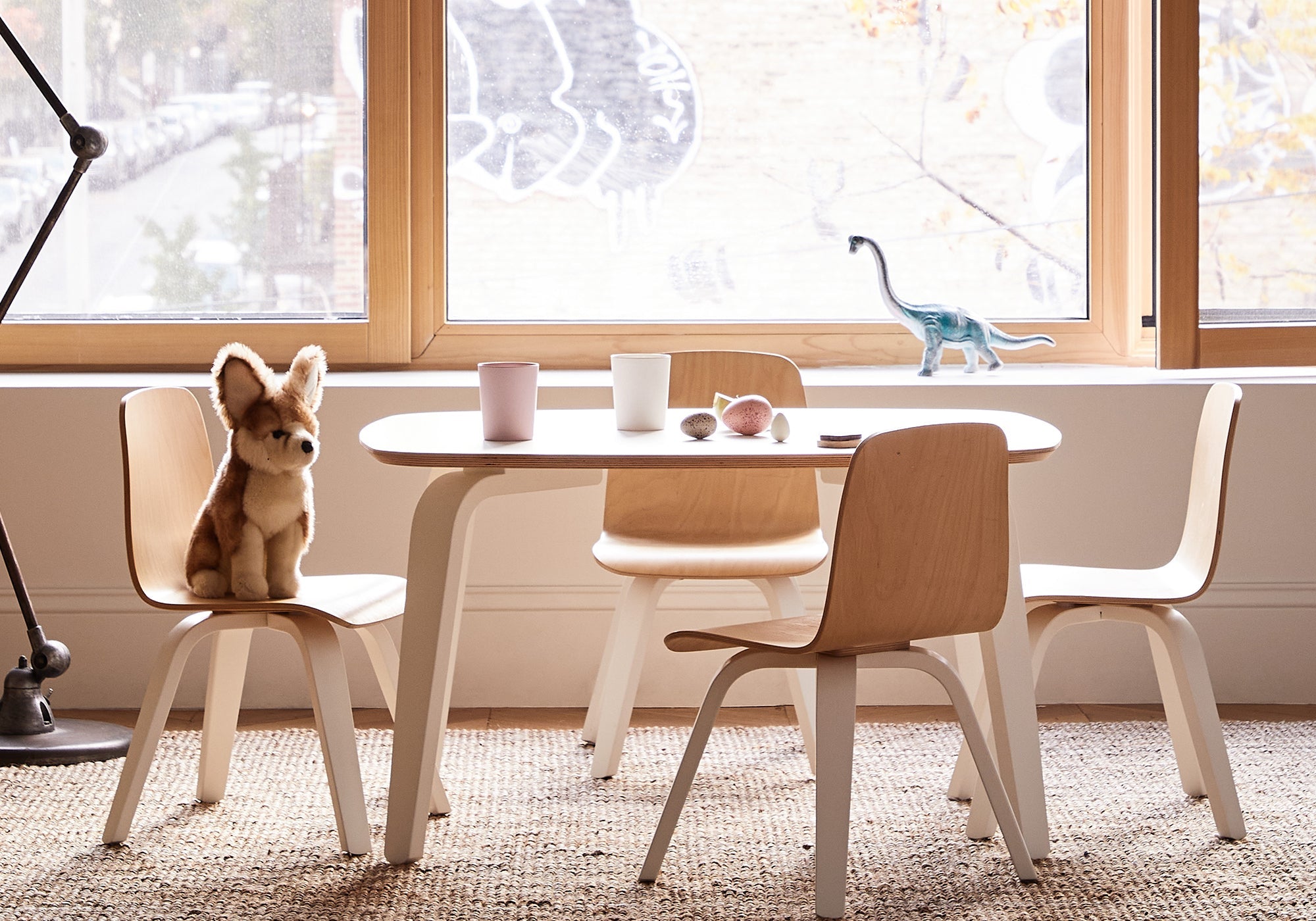 Children’s table with three chairs, plush fox toy, cups, toy fruit, and a dinosaur figurine by a window.