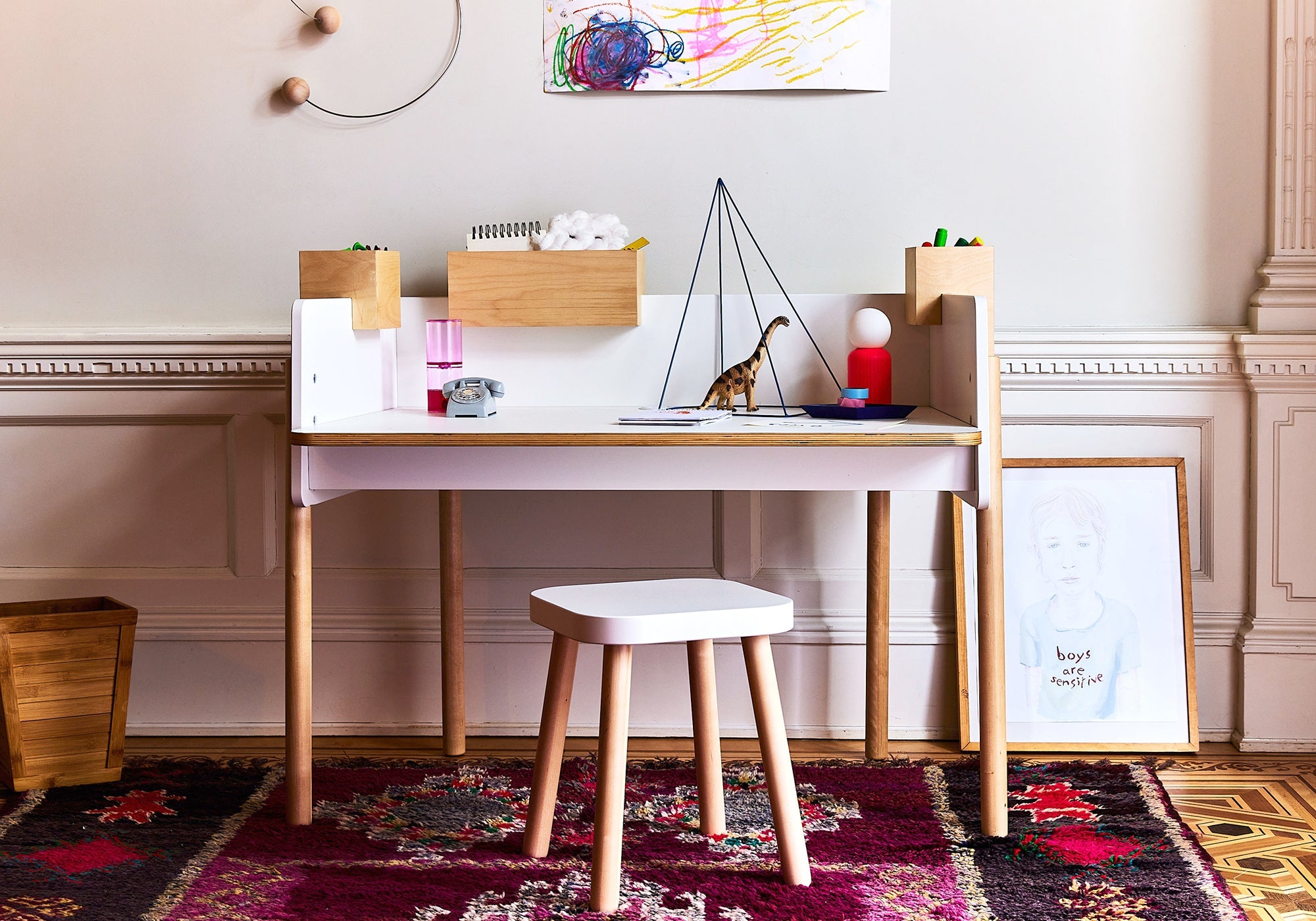 Children’s desk with art supplies, dinosaur toy, stool, and drawing on a wall above a colorful rug.