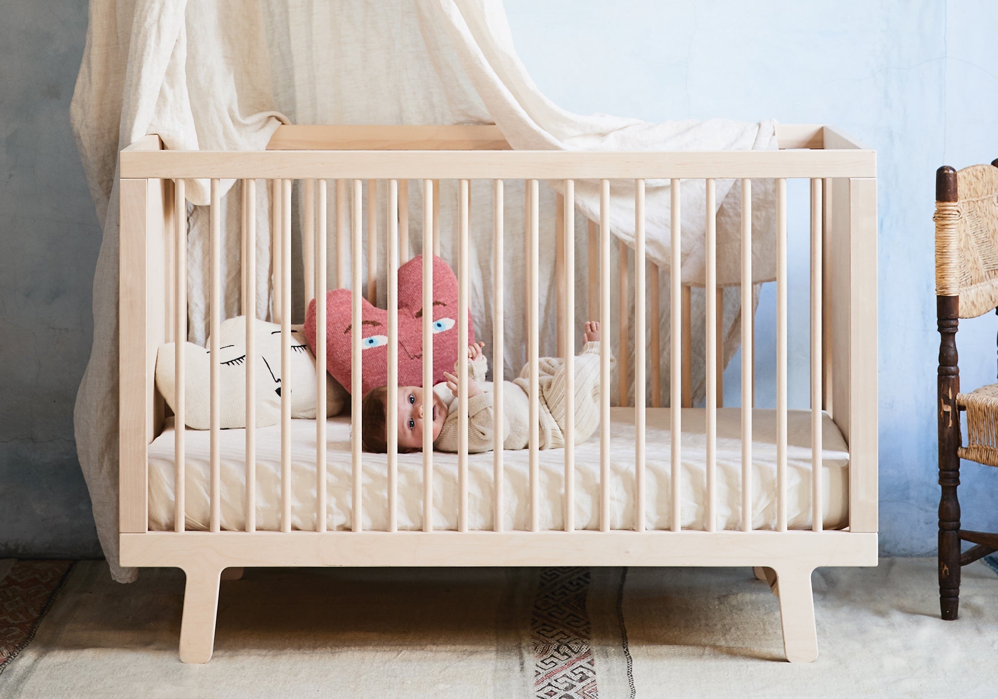 A baby lies in a wooden crib with two face-shaped pillows, next to a draped cloth and a woven chair.