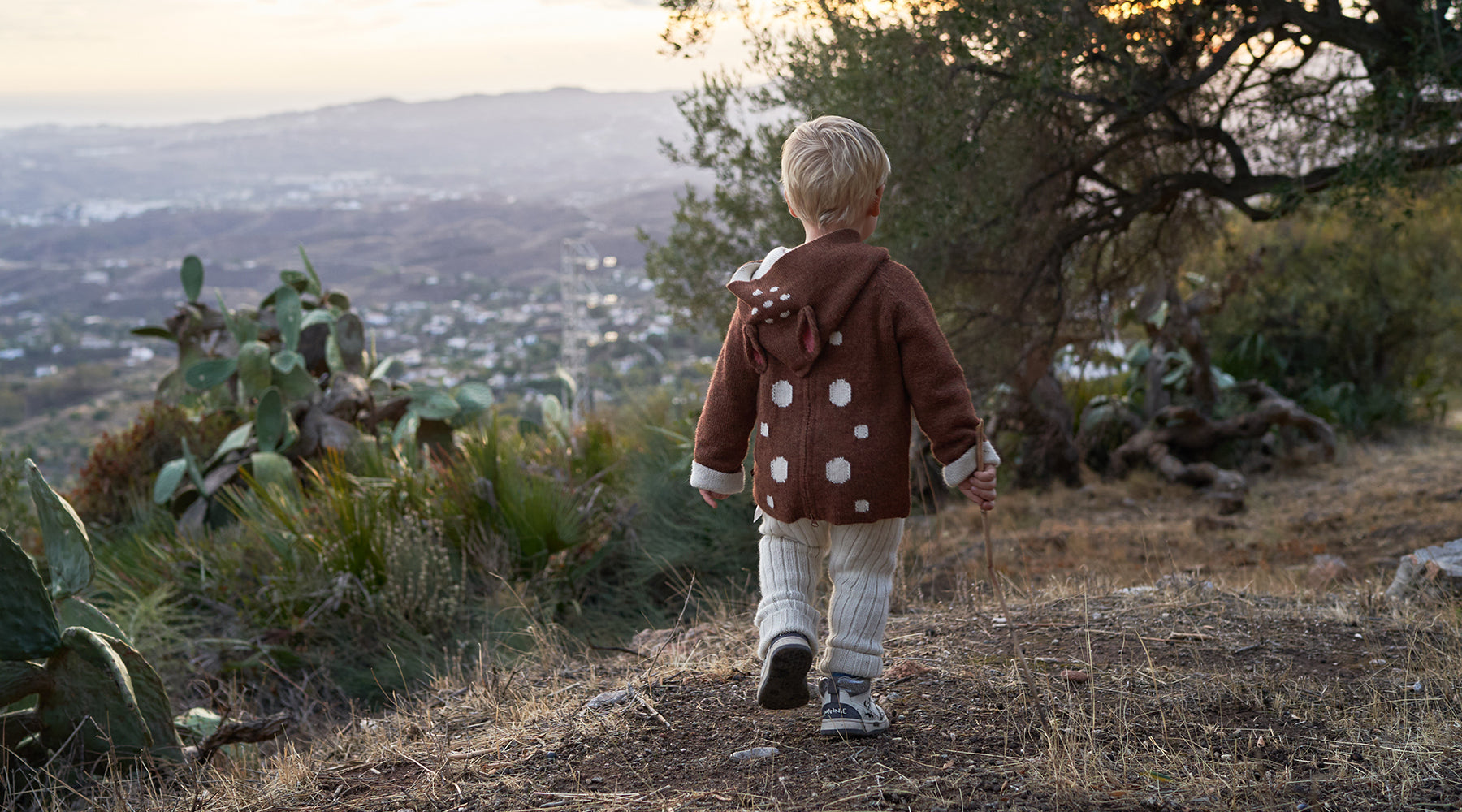 A young child in a brown hoodie walks on a dirt path with hills and a town in the distance.