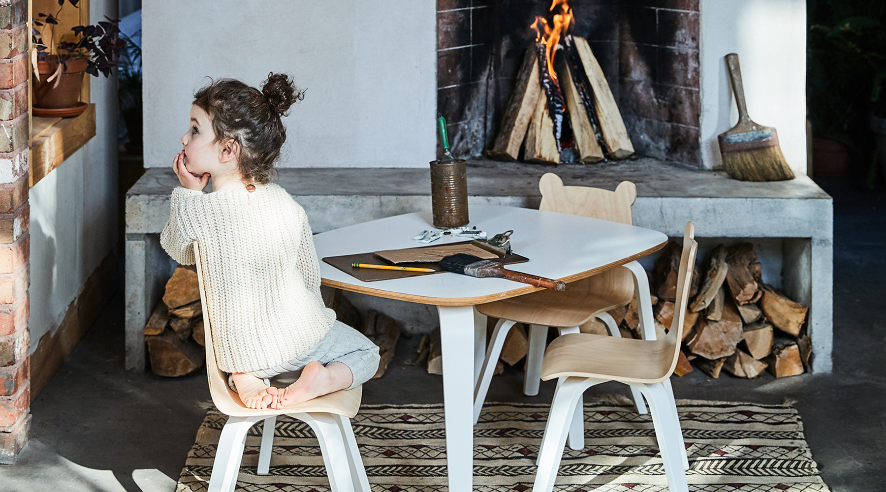 A child sits barefoot on a chair by a table, looking away, near a lit fireplace with firewood.