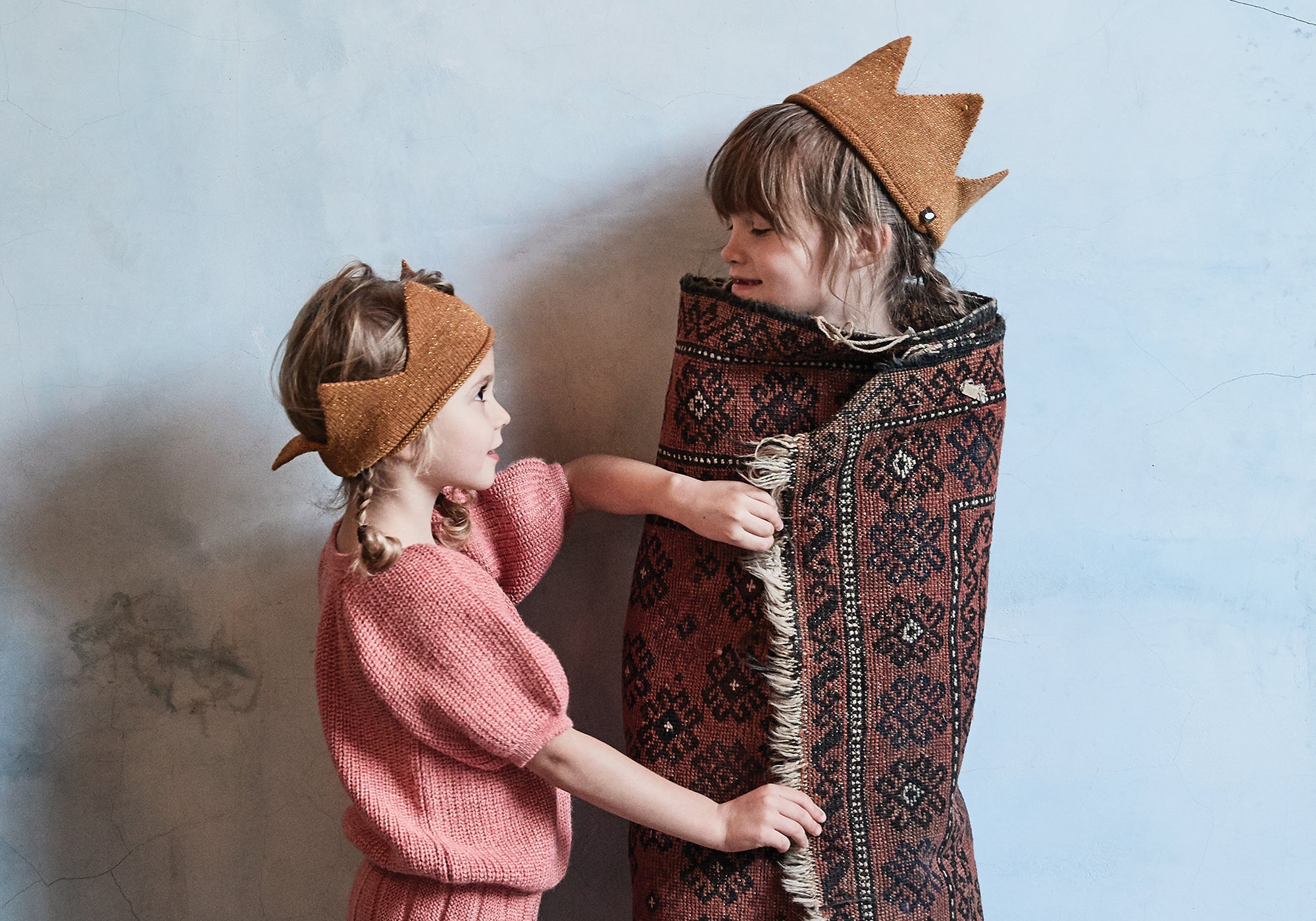 Two children play; one wears a crown and is wrapped in a patterned rug, the other wears a headscarf and pink outfit.
