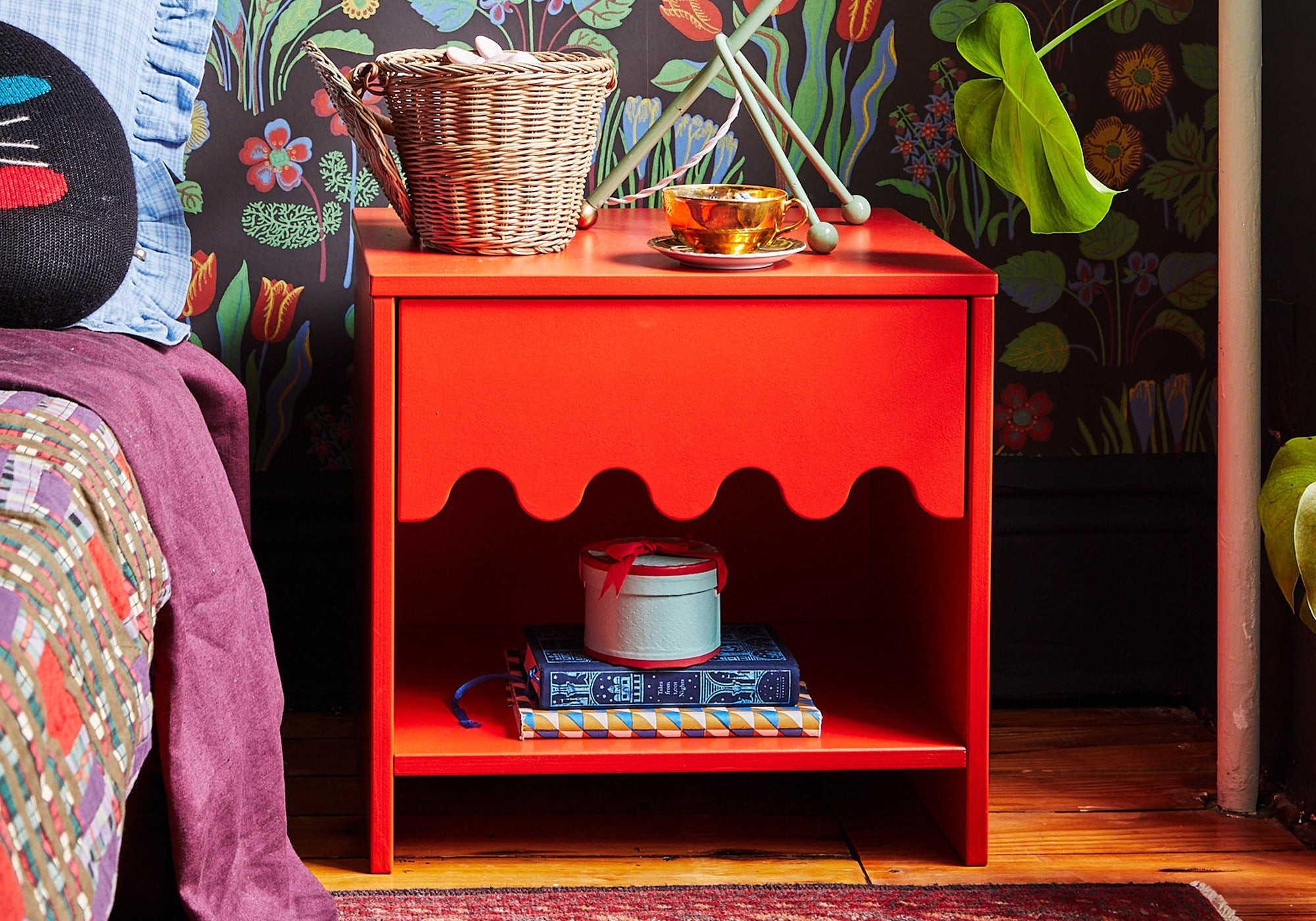 A bright red nightstand with books, a basket, and teacups, set against a floral wallpaper background.