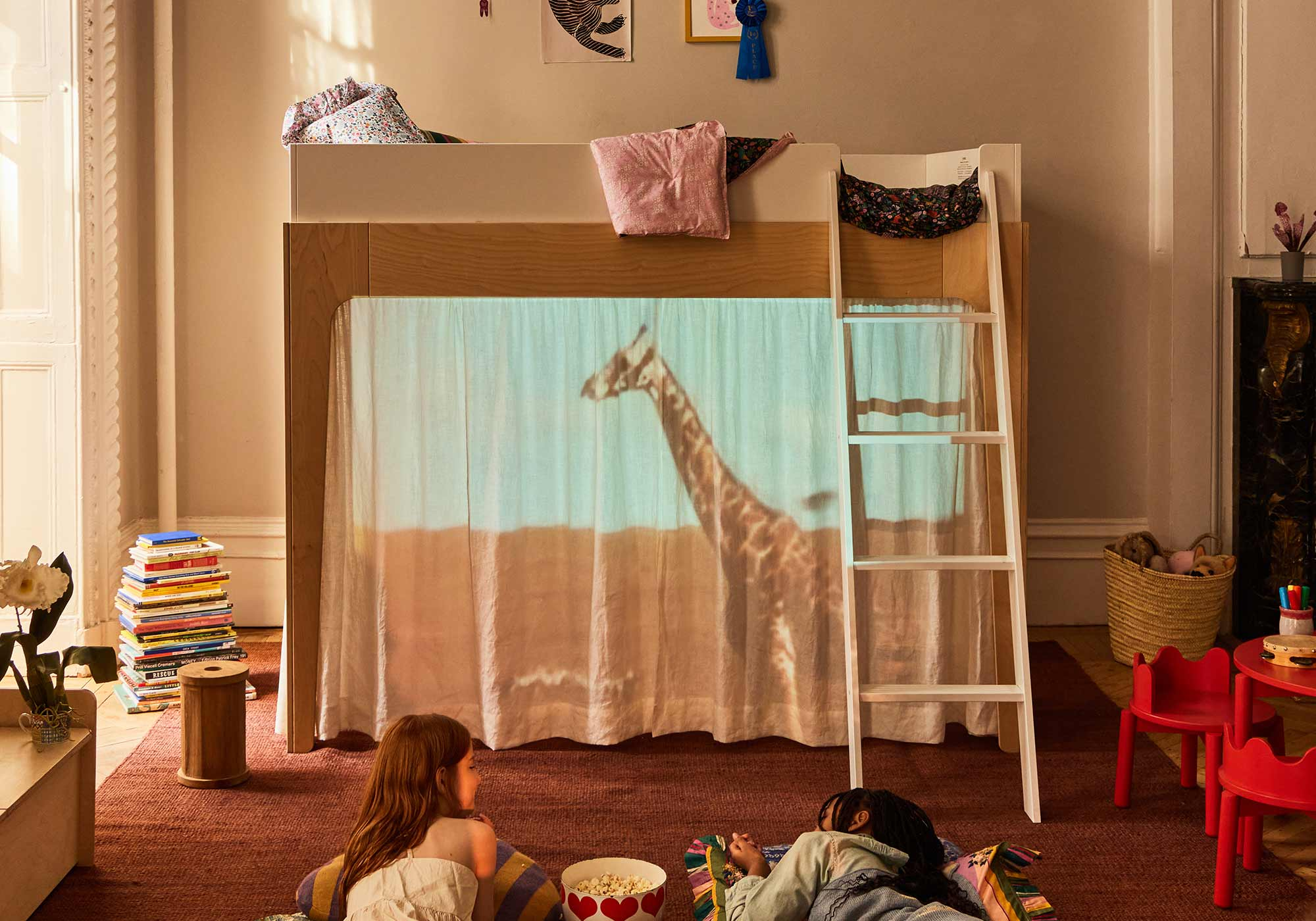 A round wooden kids' table with three chairs on a patterned rug, surrounded by toys, books, and art.