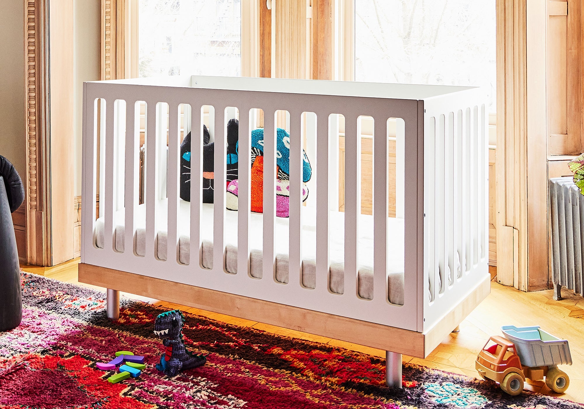 White baby crib in a sunny room with toys and stuffed animals on a colorful rug beside it.