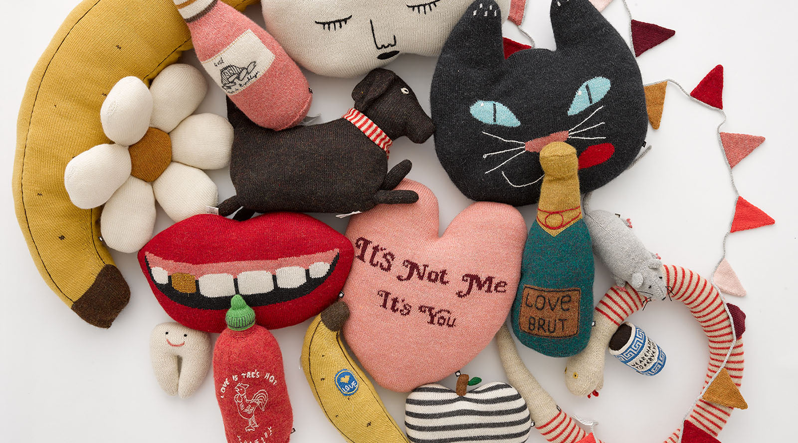 Assorted colorful plush toys, including a cat, heart, lips, bananas, and bottles, arranged on a white background.