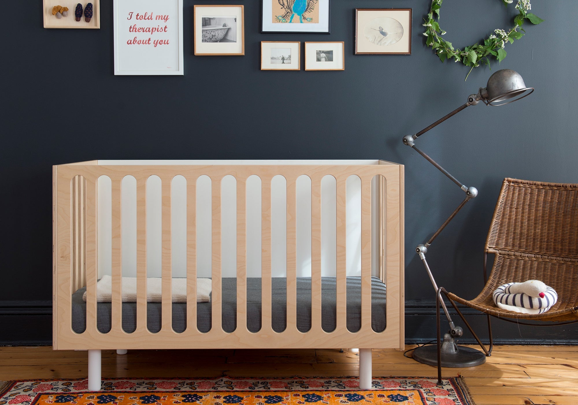 Modern baby crib in a stylish room with dark walls, wall art, a lamp, and a wicker chair.