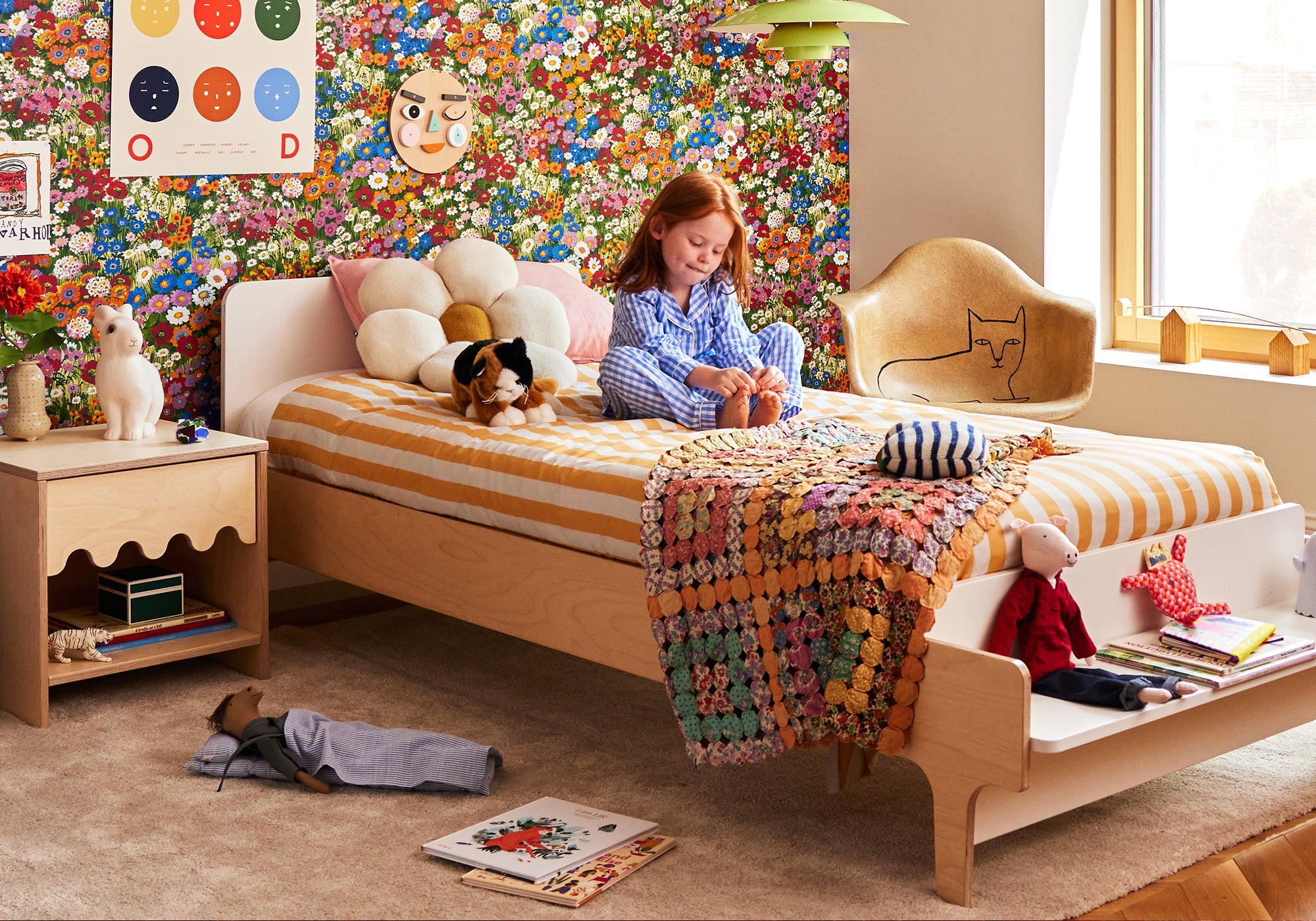 A child in pajamas sits on a bed in a colorful, toy-filled bedroom with a floral wallpaper accent wall.