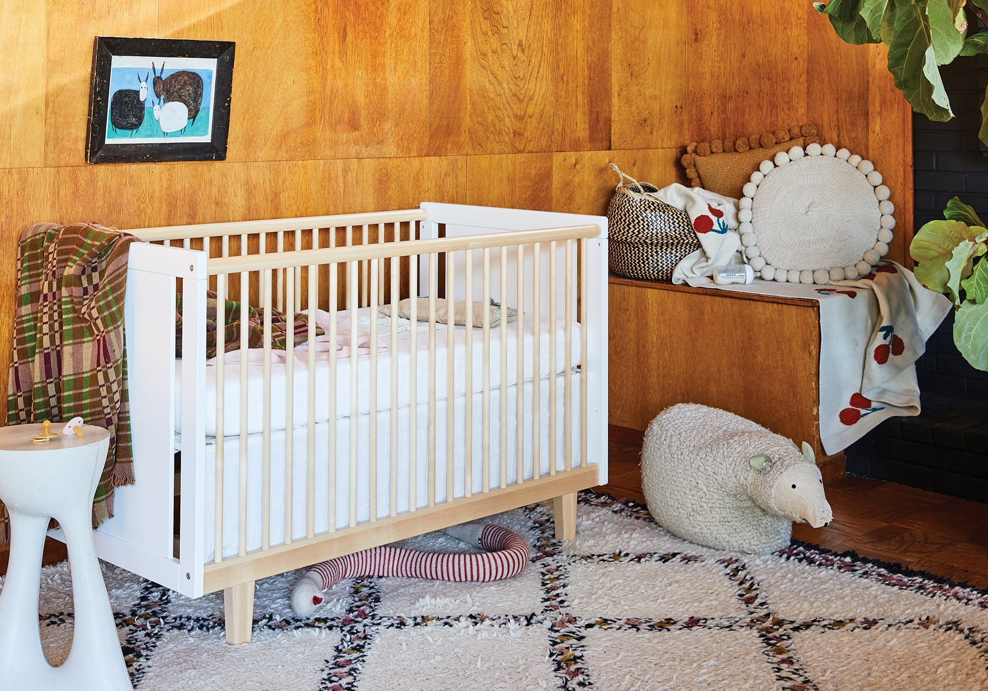 Modern nursery with a white crib, toys, pillows, and a cozy rug on a wooden floor with warm wood wall panels.
