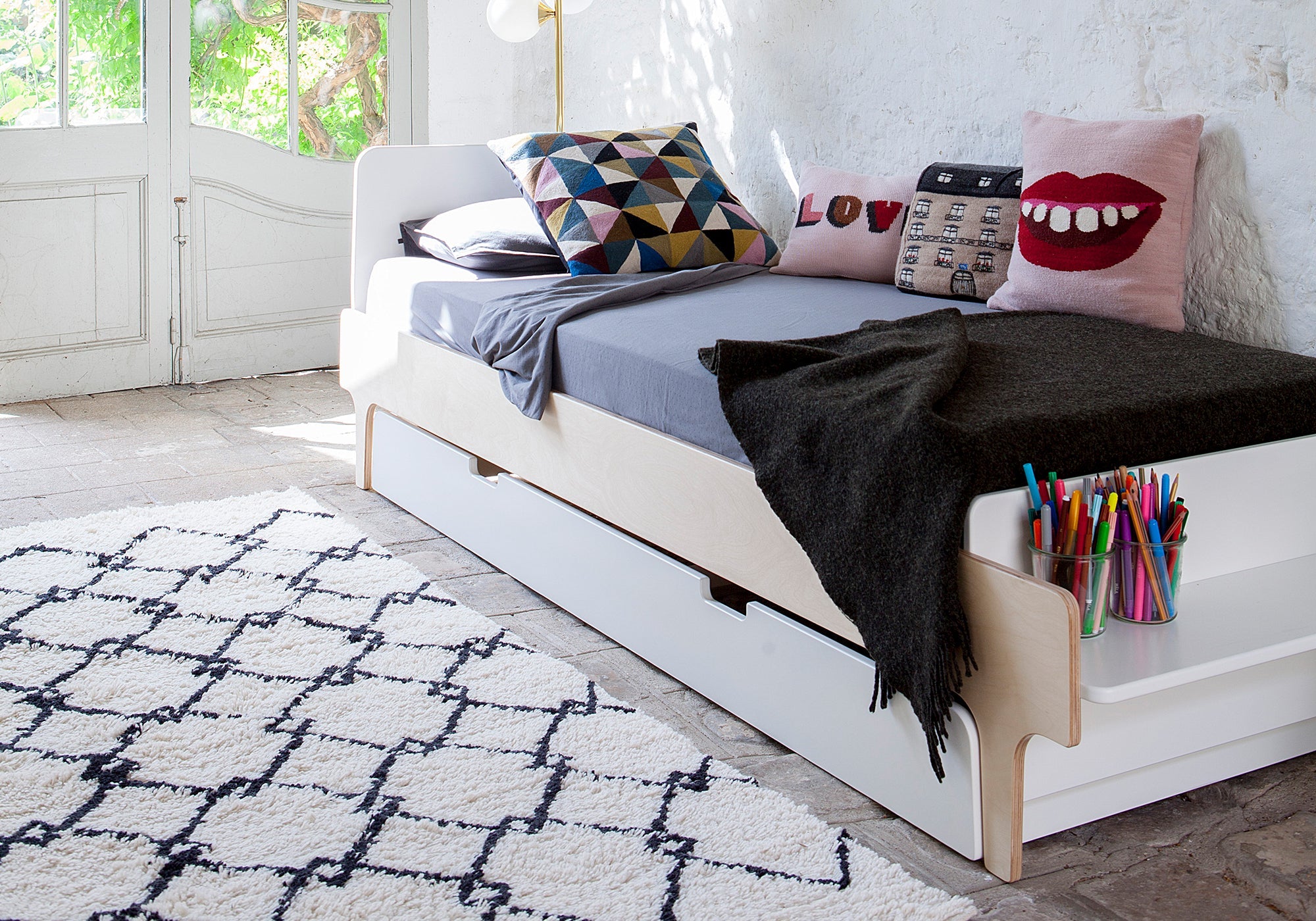 A stylish bed with geometric and lip print pillows, a cozy blanket, and colored pencils in a bright, airy room.