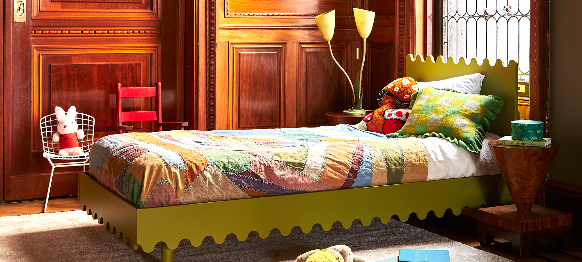 Colorful child’s bedroom with a wavy-edged green bed, patchwork quilt, plush toys, and wooden panel walls.