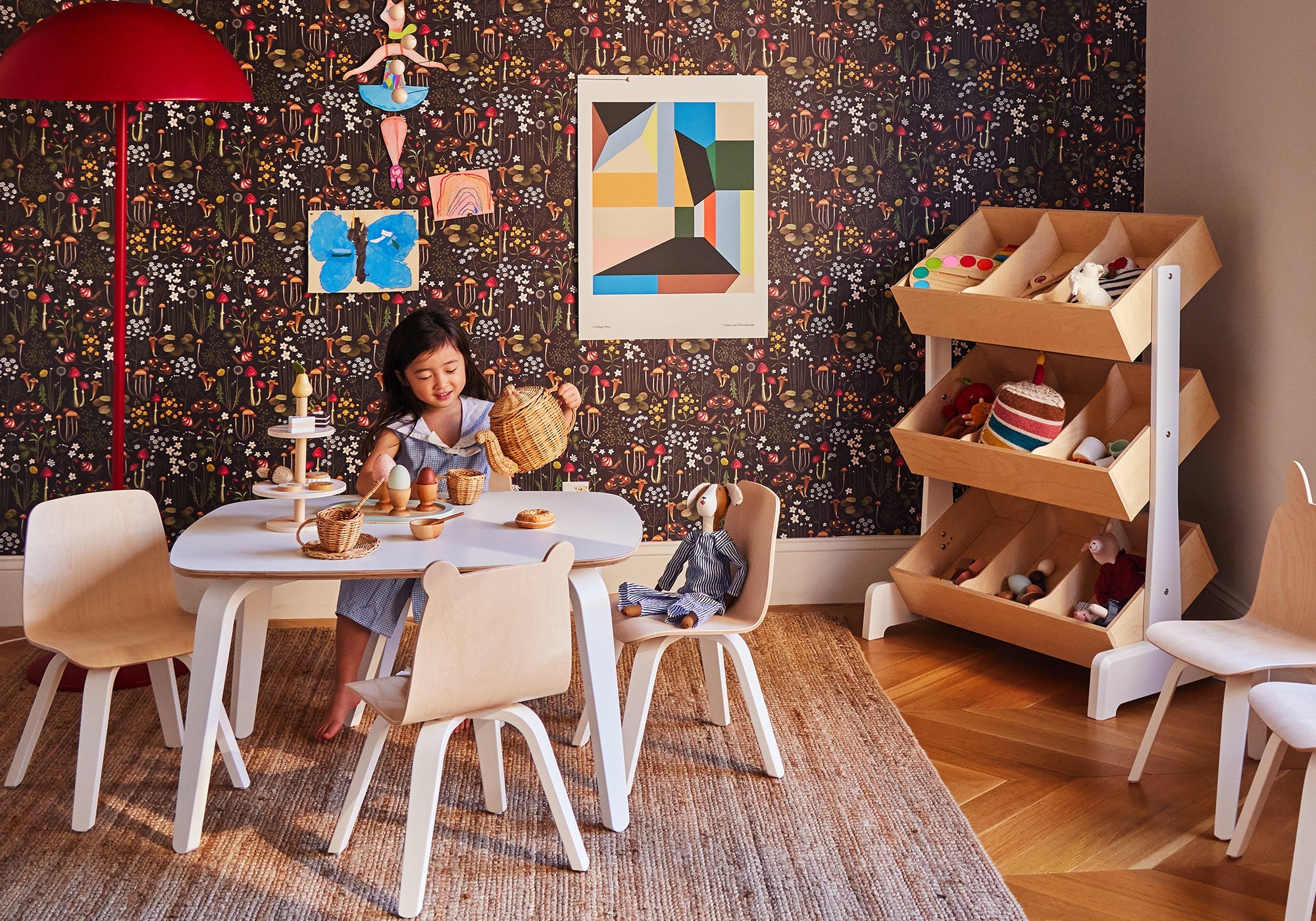 A young girl plays tea party at a small table in a colorful, modern children's playroom with toys and art.