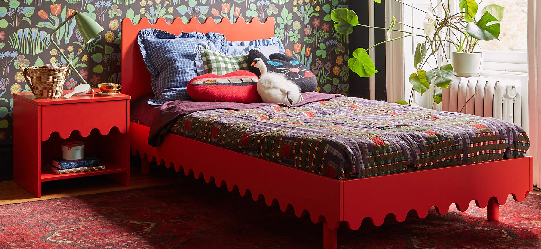 Bright red bed and nightstand in a colorful room with patterned bedding, pillows, and leafy wallpaper.