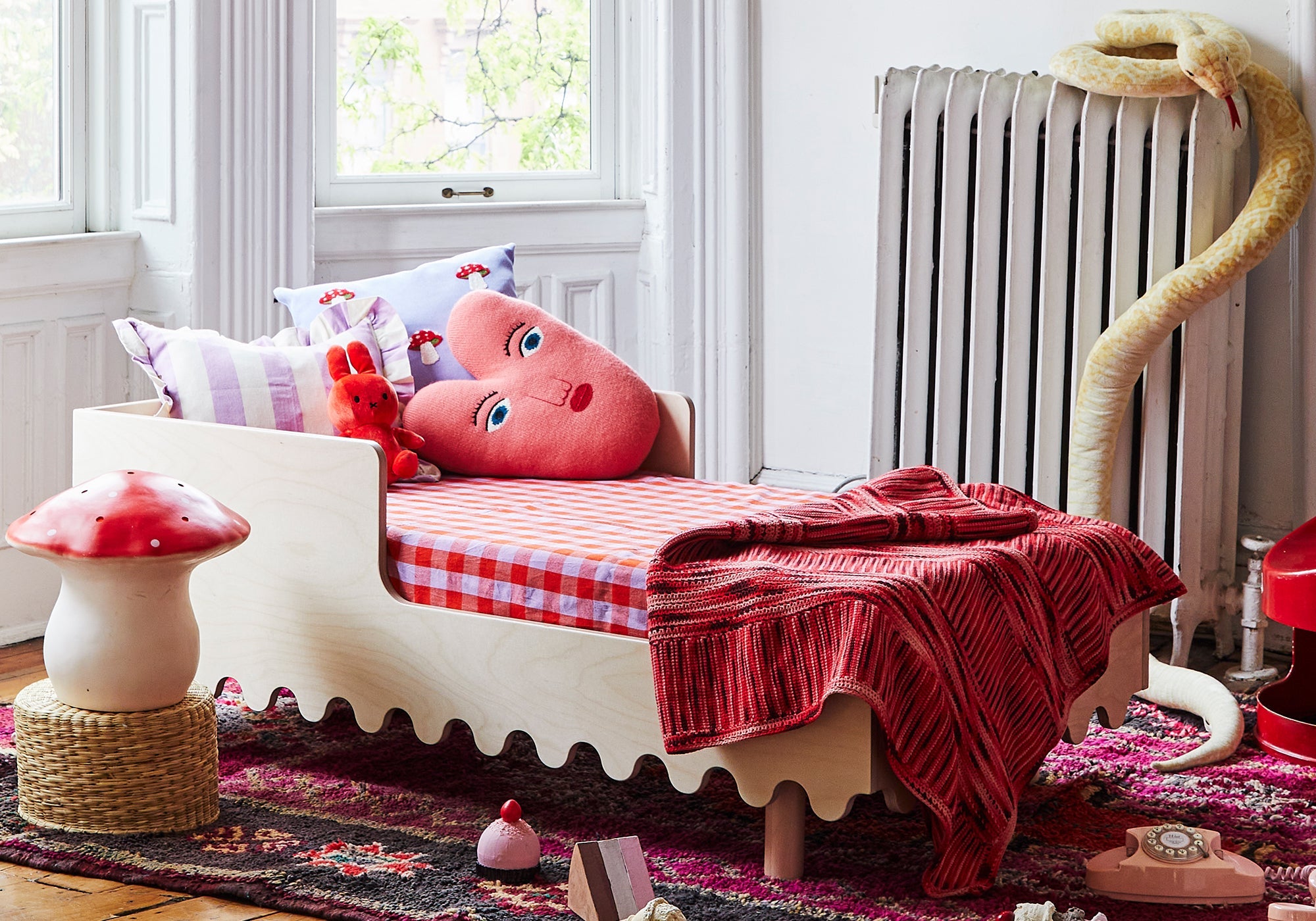 Children’s bed with a heart pillow, red blanket, stuffed animals, and a large snake near a radiator.