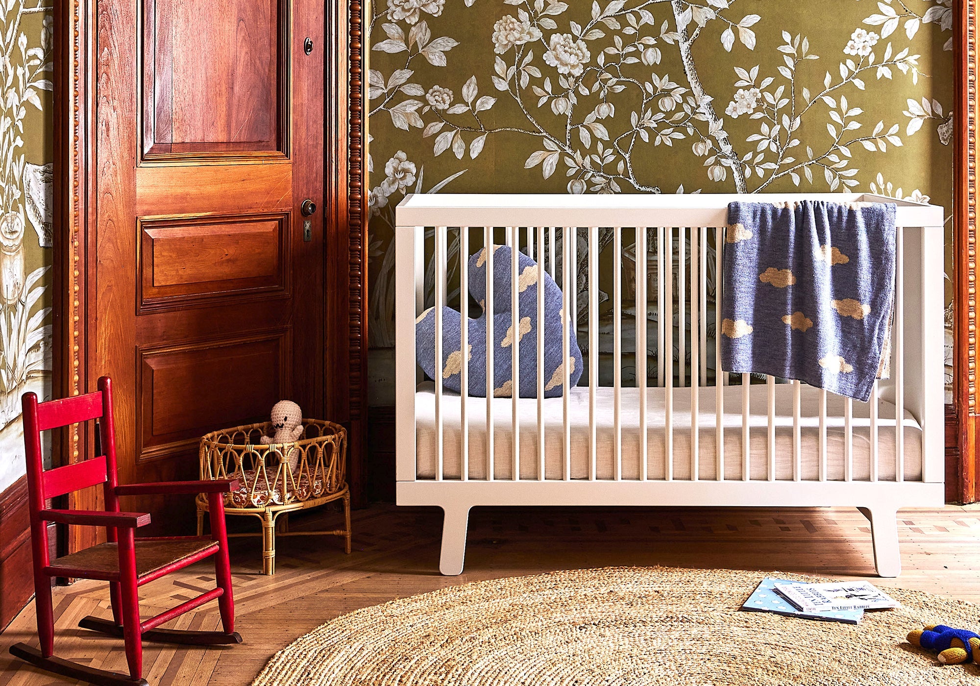 Nursery with a white crib, blue blanket, red chair, and floral wallpaper.