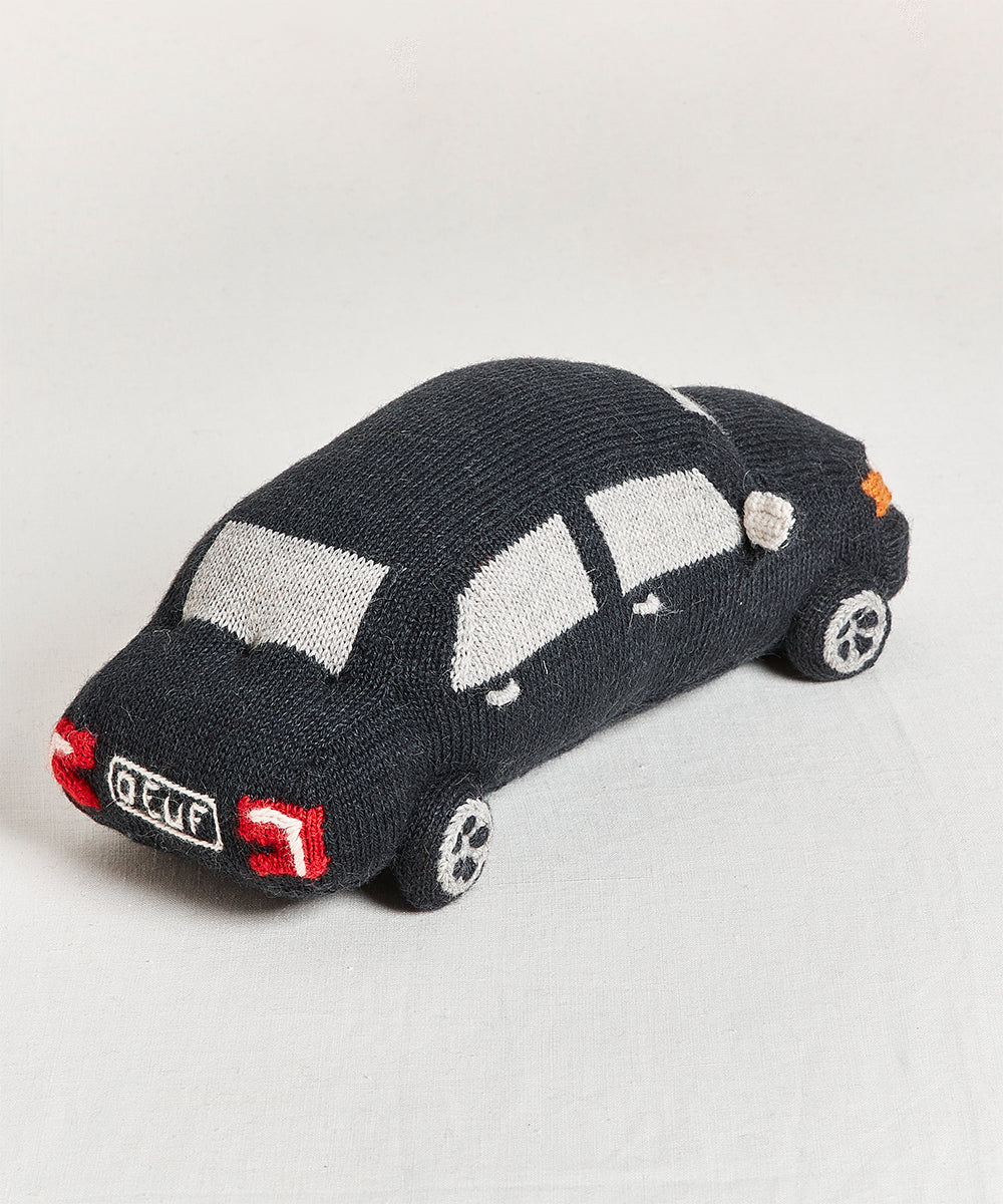 The My Ride Pillow is a knitted black toy car featuring white windows and red tail lights, set on a gray surface.