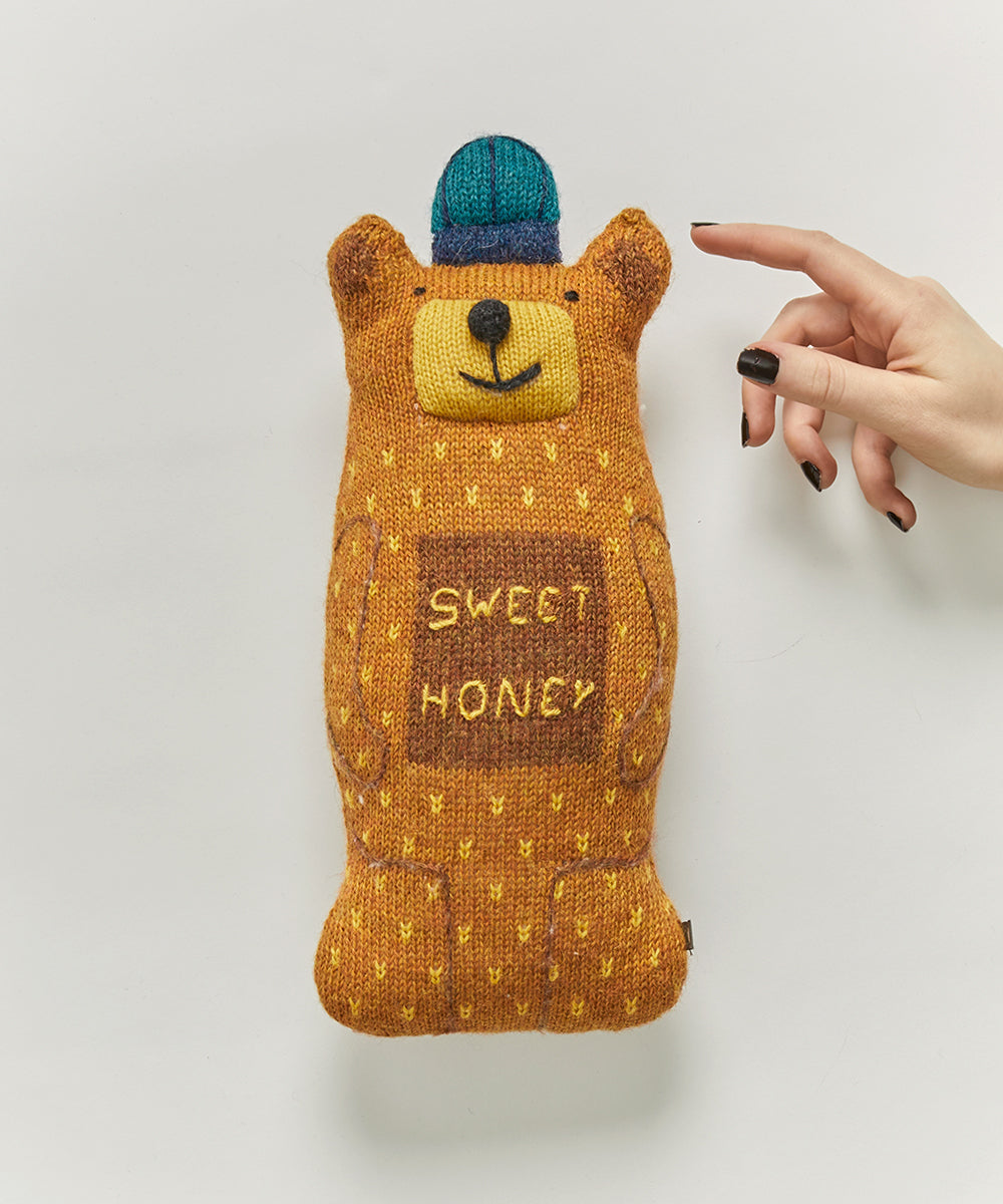 A hand with black nails touches the Honey Bear Pillow, a brown plush bear with "Sweet Honey" and a blue cap stitched on it.