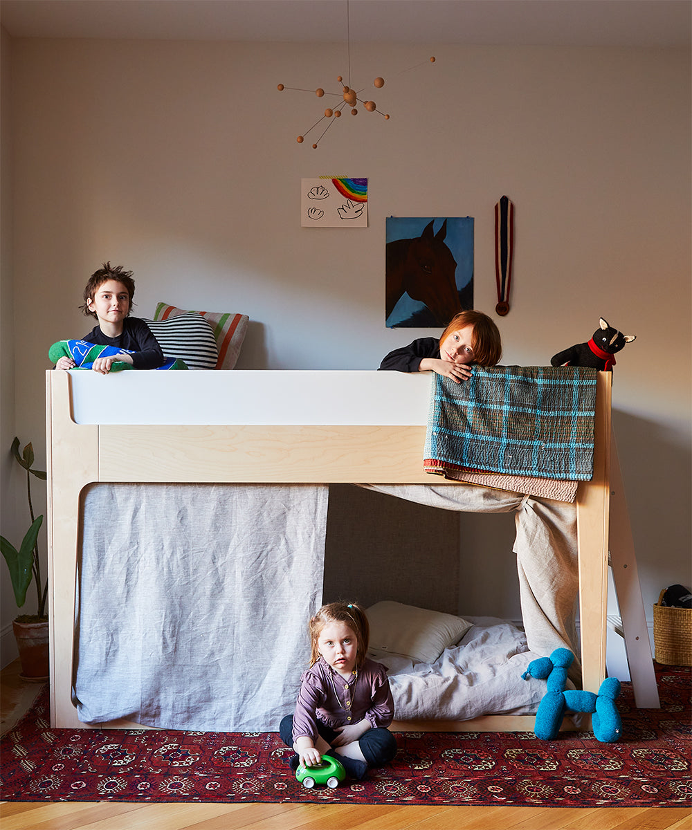 Two kids enjoy the Perch Nest Bed loft above a child playing beneath in a cozy room featuring a red rug and playful decor.