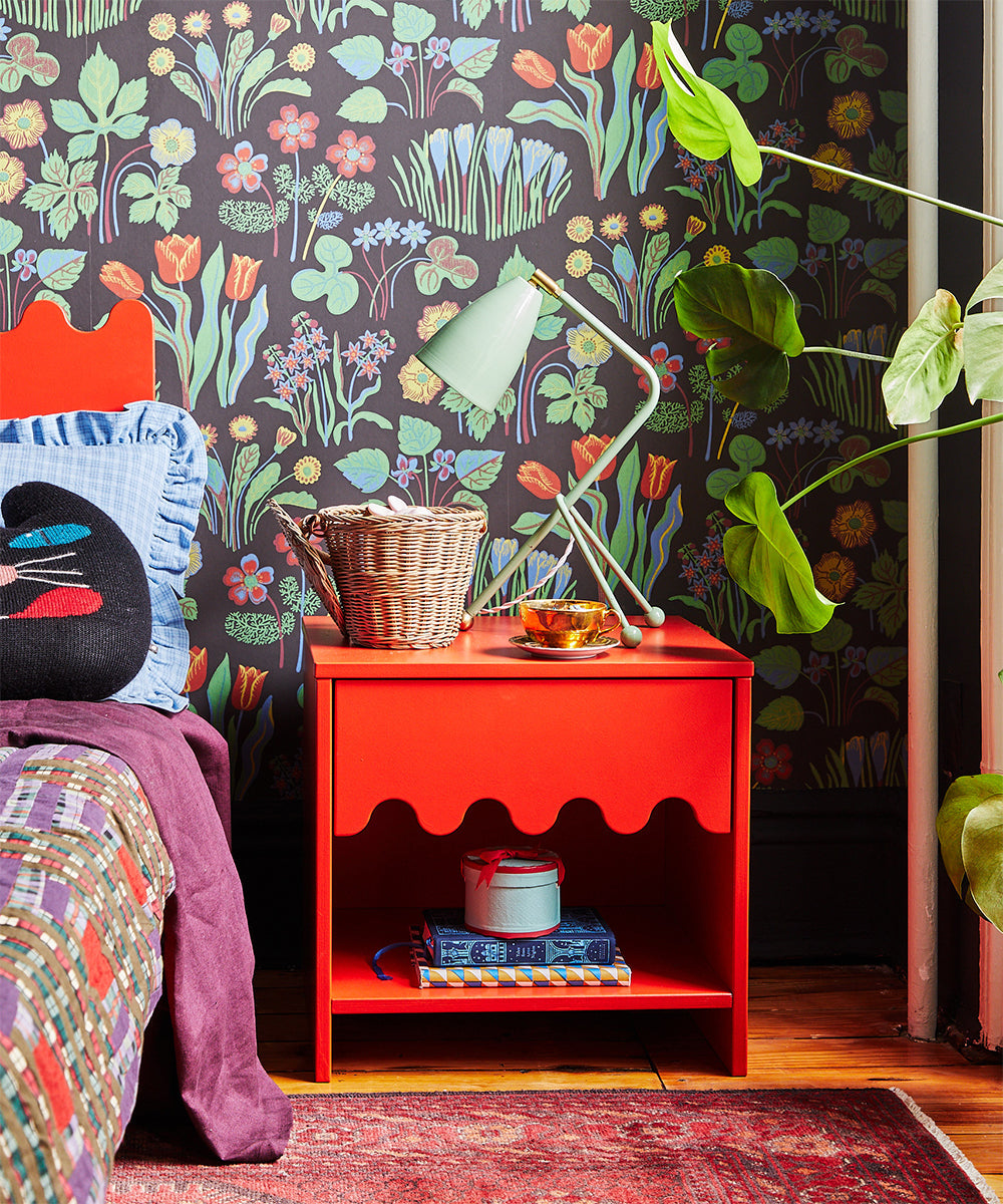 A colorful bedroom featuring a Moss Nightstand, a green lamp, floral wallpaper, and plants. A basket and snacks rest on the table.