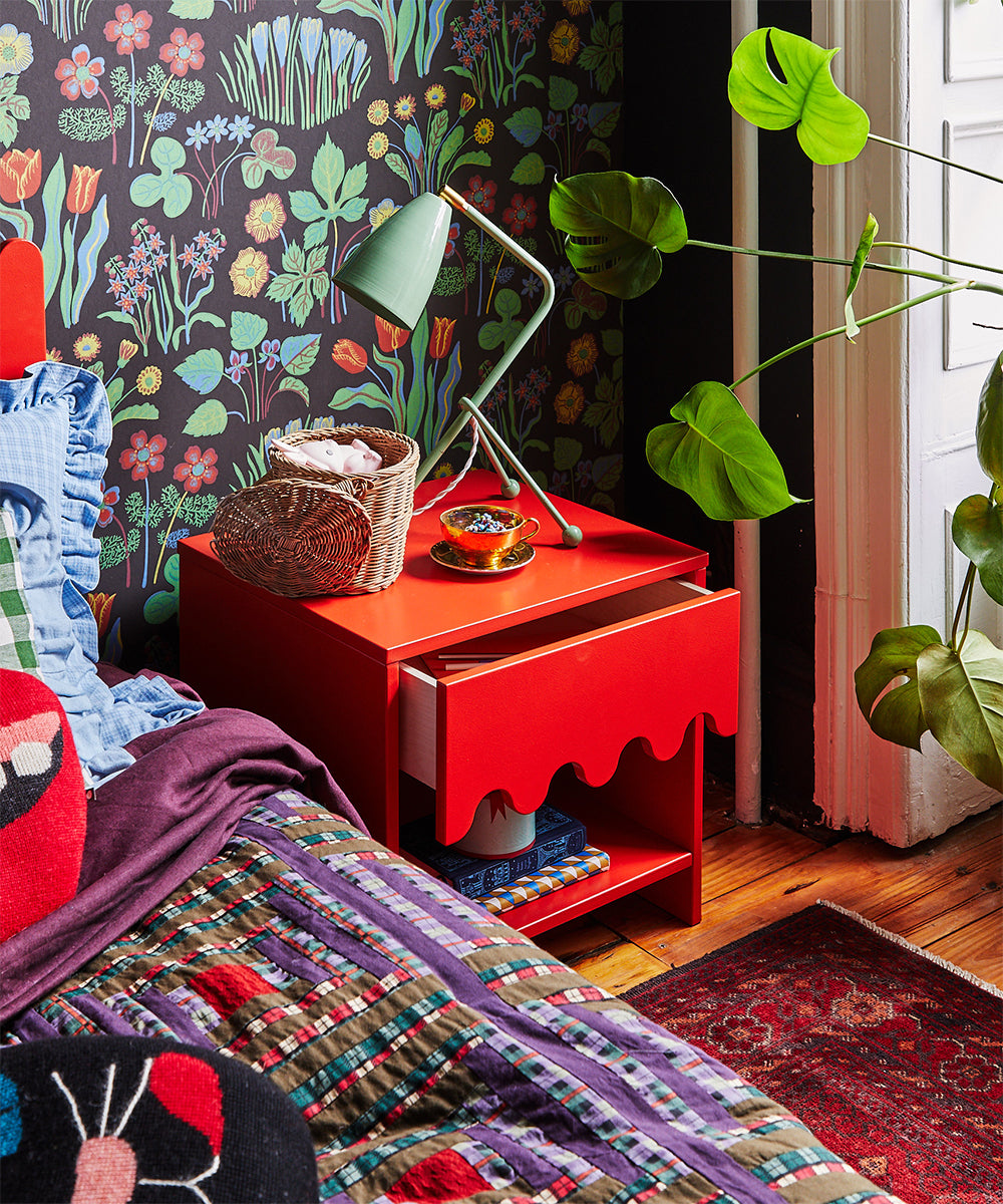A colorful bedroom featuring floral wallpaper, a Moss Nightstand, a green lamp, and a bed with vibrant patterned bedding.