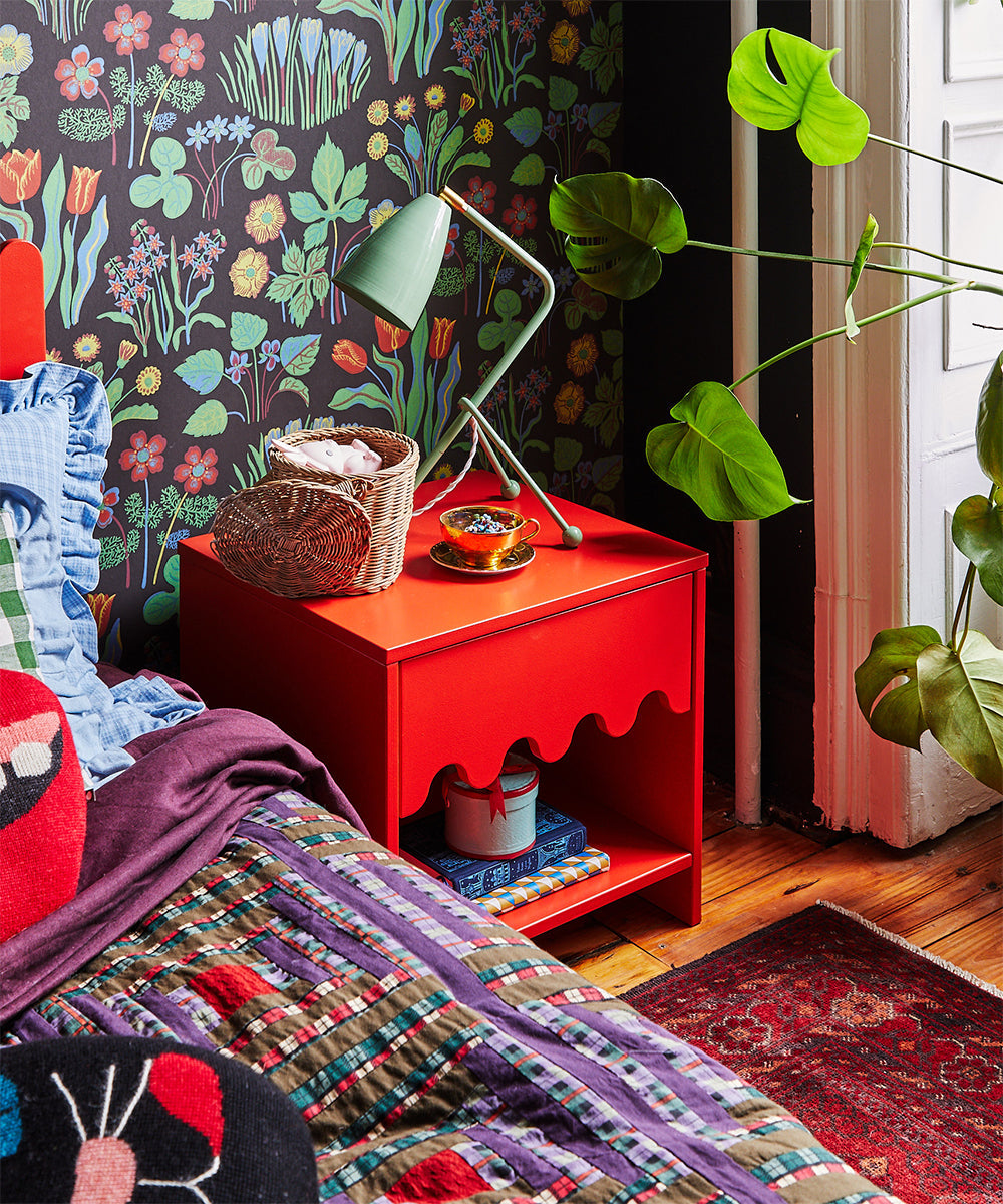 A vibrant bedroom featuring a Moss Nightstand, a green lamp, floral wallpaper, colorful bedding, and plants adds charm.