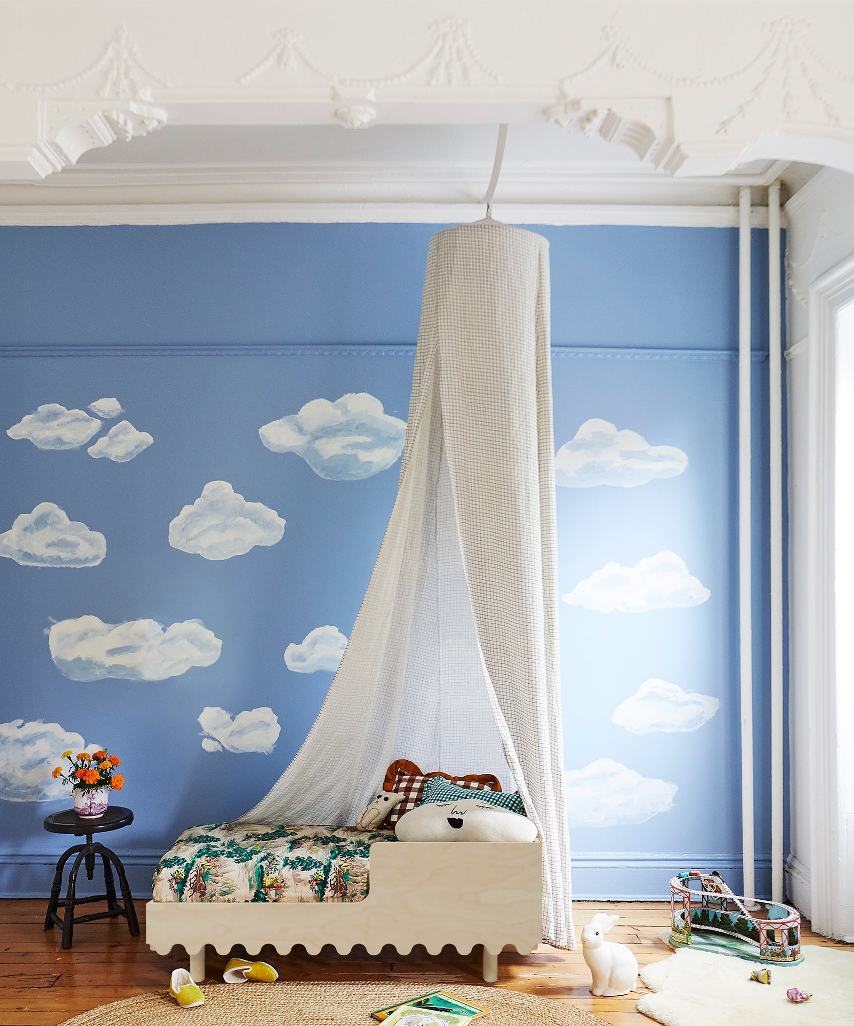 The Moss Toddler Bed with canopy is featured in a blue room decorated with painted clouds, books, and toys scattered on the floor.