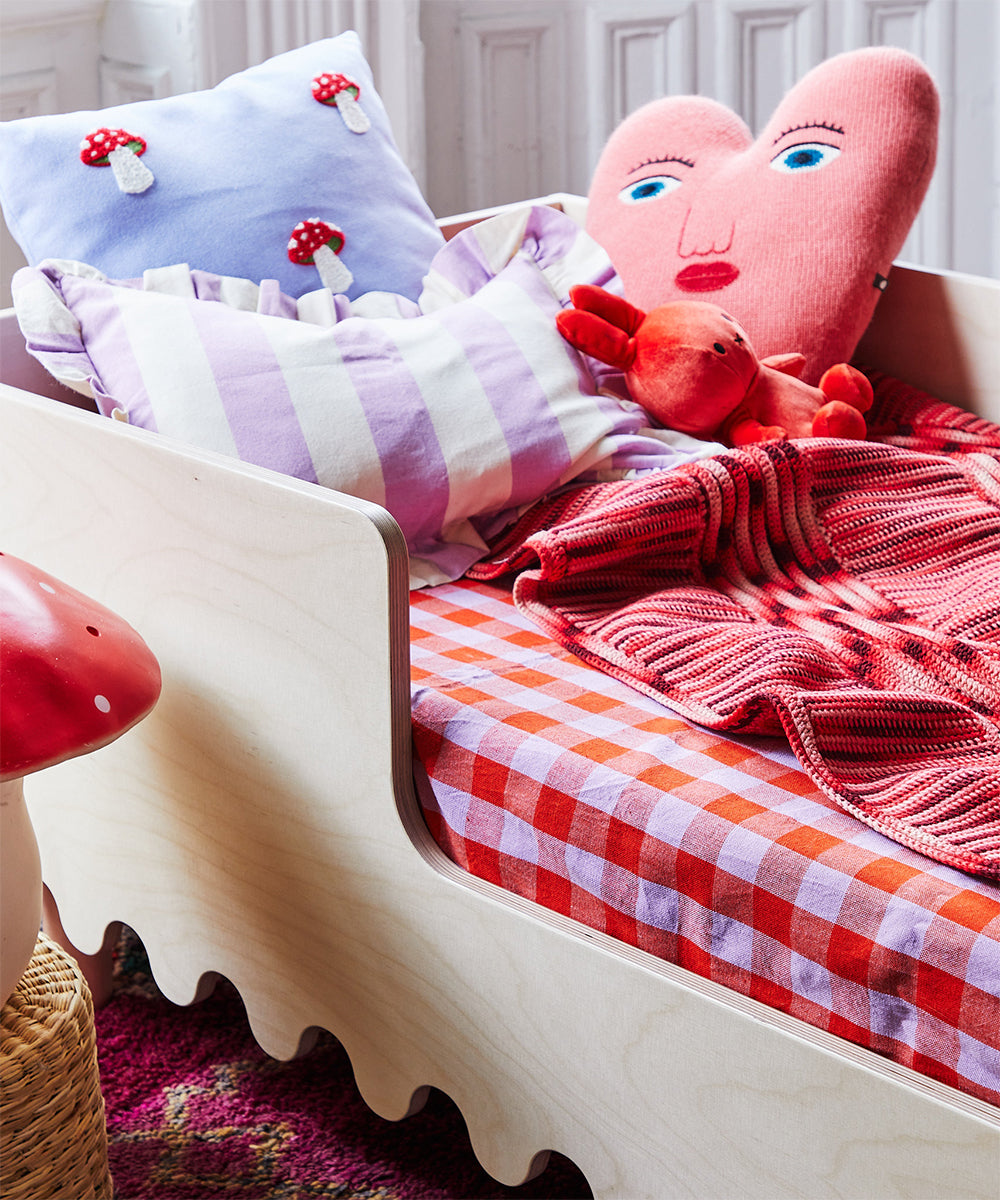 The Moss Toddler Bed is adorned with heart and mushroom pillows, checkered bedding, and a red knitted blanket for a cozy feel.