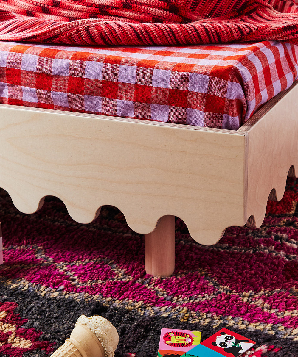 The Moss Toddler Bed features wooden construction with wavy edges, adorned with red checkered sheets and placed on a colorful carpet surrounded by toys.