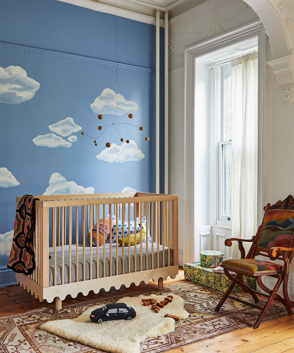 A nursery featuring the Moss Crib, a cloud-themed wall design, a cozy chair, a soft rug, and a hanging mobile adorned with wooden balls.