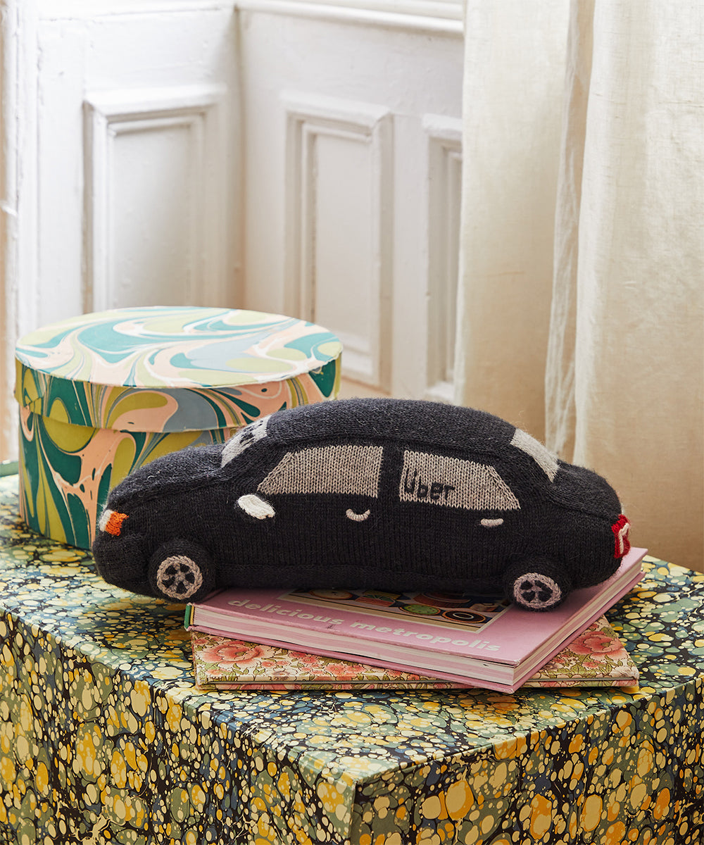 The My Ride Pillow, a black knitted car toy, sits on colorful books and a box on a marbled-patterned table near a window.