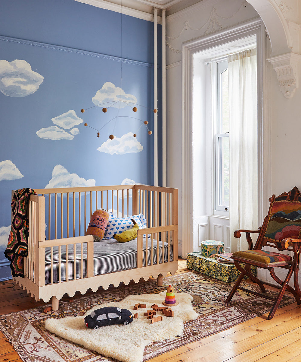 A cozy nursery features the Moss Crib Conversion Kit, a cloud mural, toys on a rug, and natural light streaming through a window.