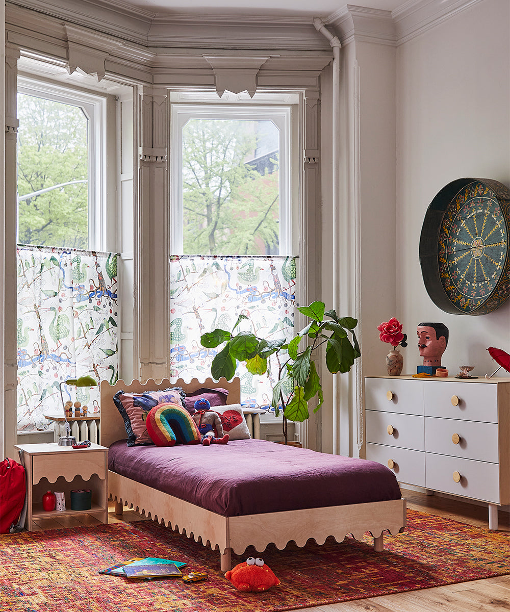 A colorful bedroom featuring a purple bed, decorative pillows, a leafy plant, artwork, patterned curtains, and complemented by the Moss Nightstand and dresser.