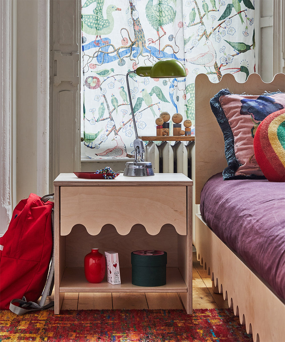 The Moss Nightstand adorned with red items, books, and a lamp stands next to a bed and backpack, with vibrant curtains as the backdrop.