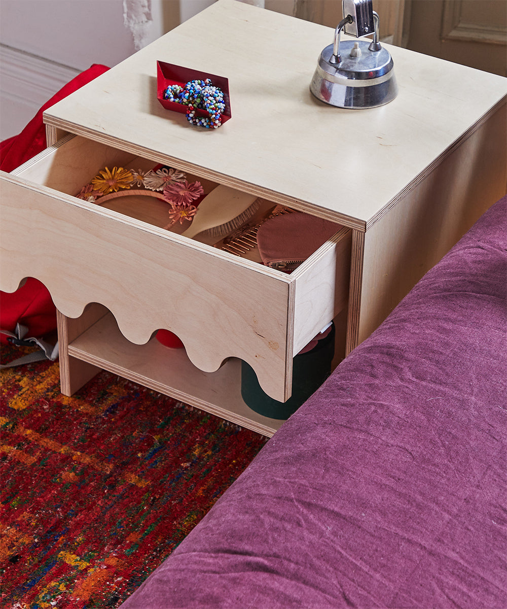 The Moss Nightstand, featuring a wavy-edged open drawer filled with accessories, sits on a colorful rug beside a purple bedspread.