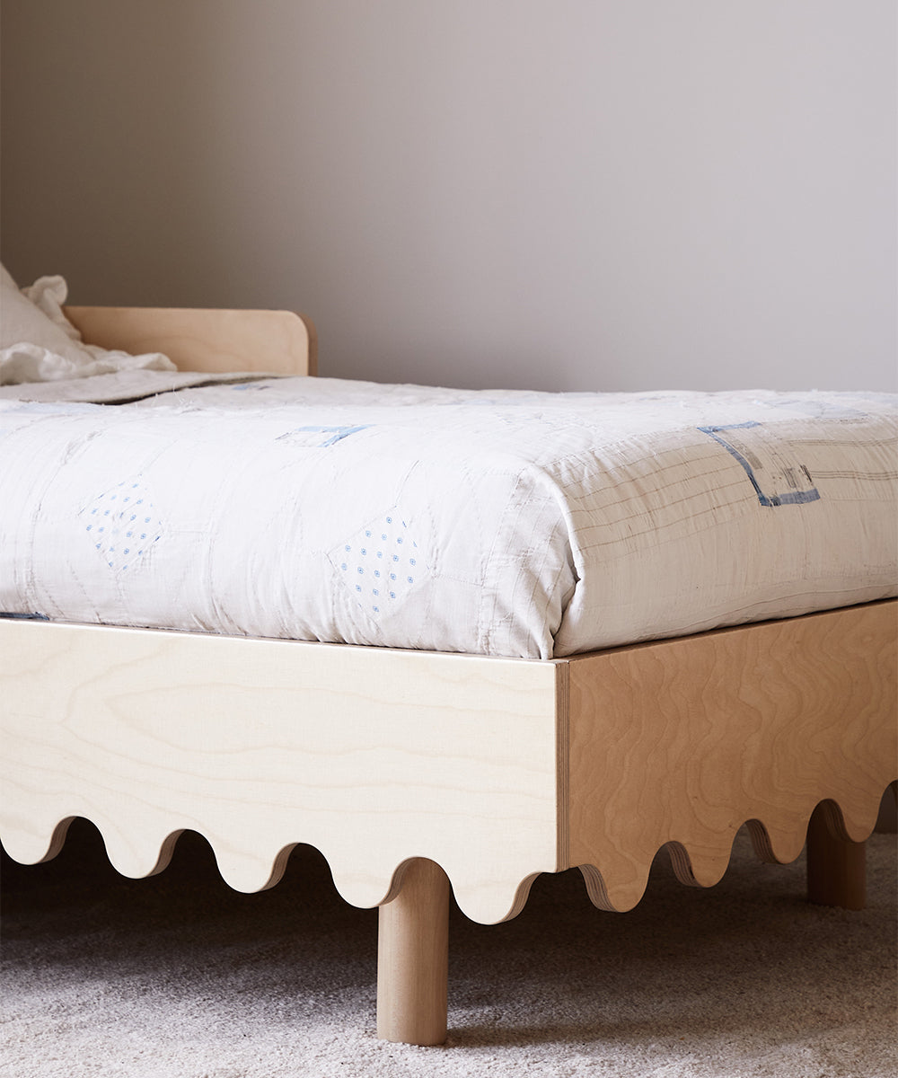 The Moss Toddler Bed features a minimalist wooden design with a wavy-edged frame, light quilted bedding, and is set against a plain wall on beige carpet.