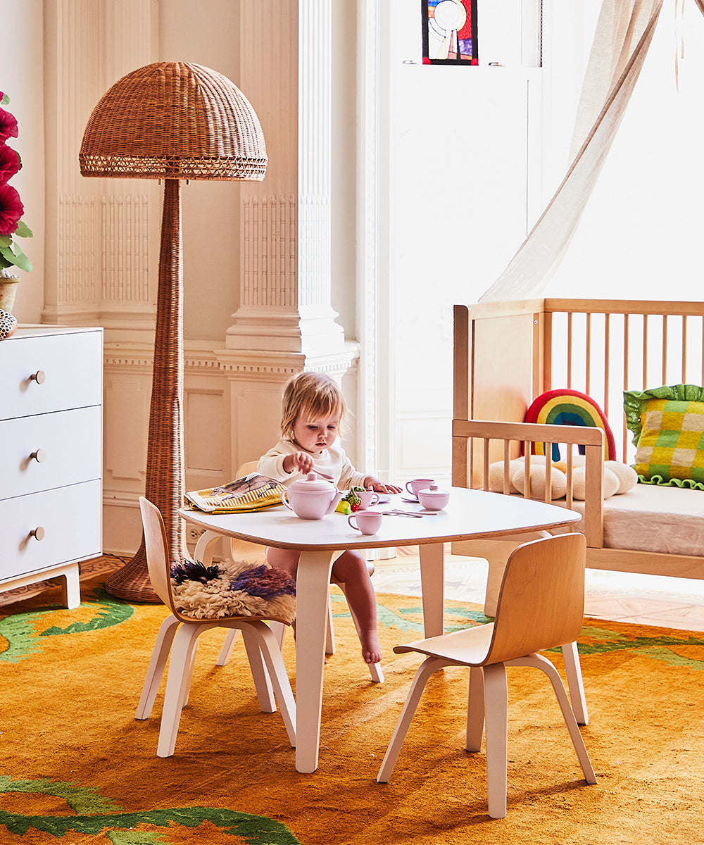 A toddler sits at a small table in a cozy room featuring a crib, Play Chair (Set of 2), and a lamp, with rugs and toys scattered around.