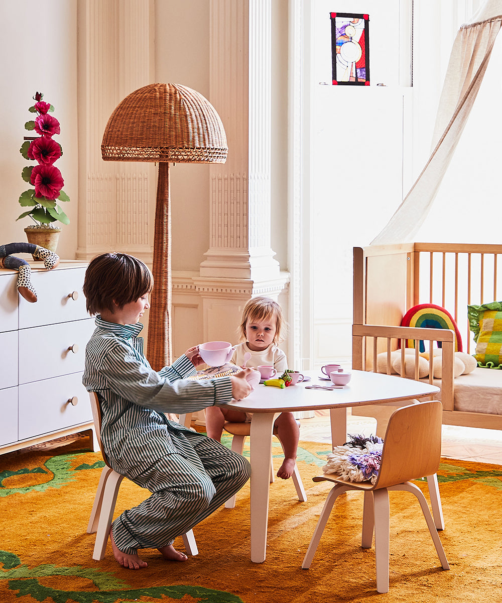 Two children enjoy a tea party on a small table, accompanied by cozy Play Chairs in a colorful room featuring a crib and tall lamp.