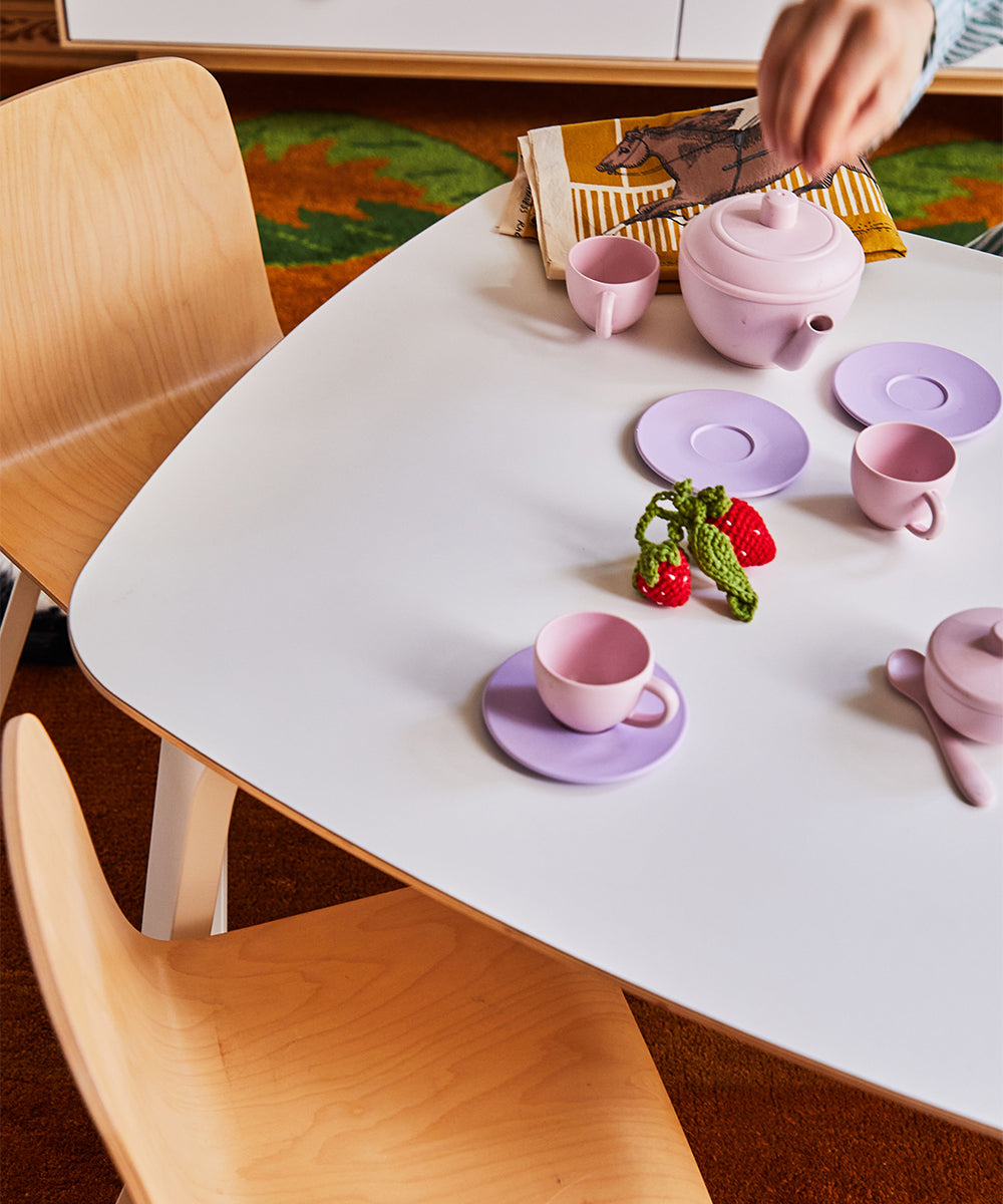 A hand reaches for a pink teapot on a play table featuring pink cups, saucers, a toy strawberry, and pea pod. Nearby, two wooden chairs rest on a brown and green rug in this child-friendly area made from sustainable materials.