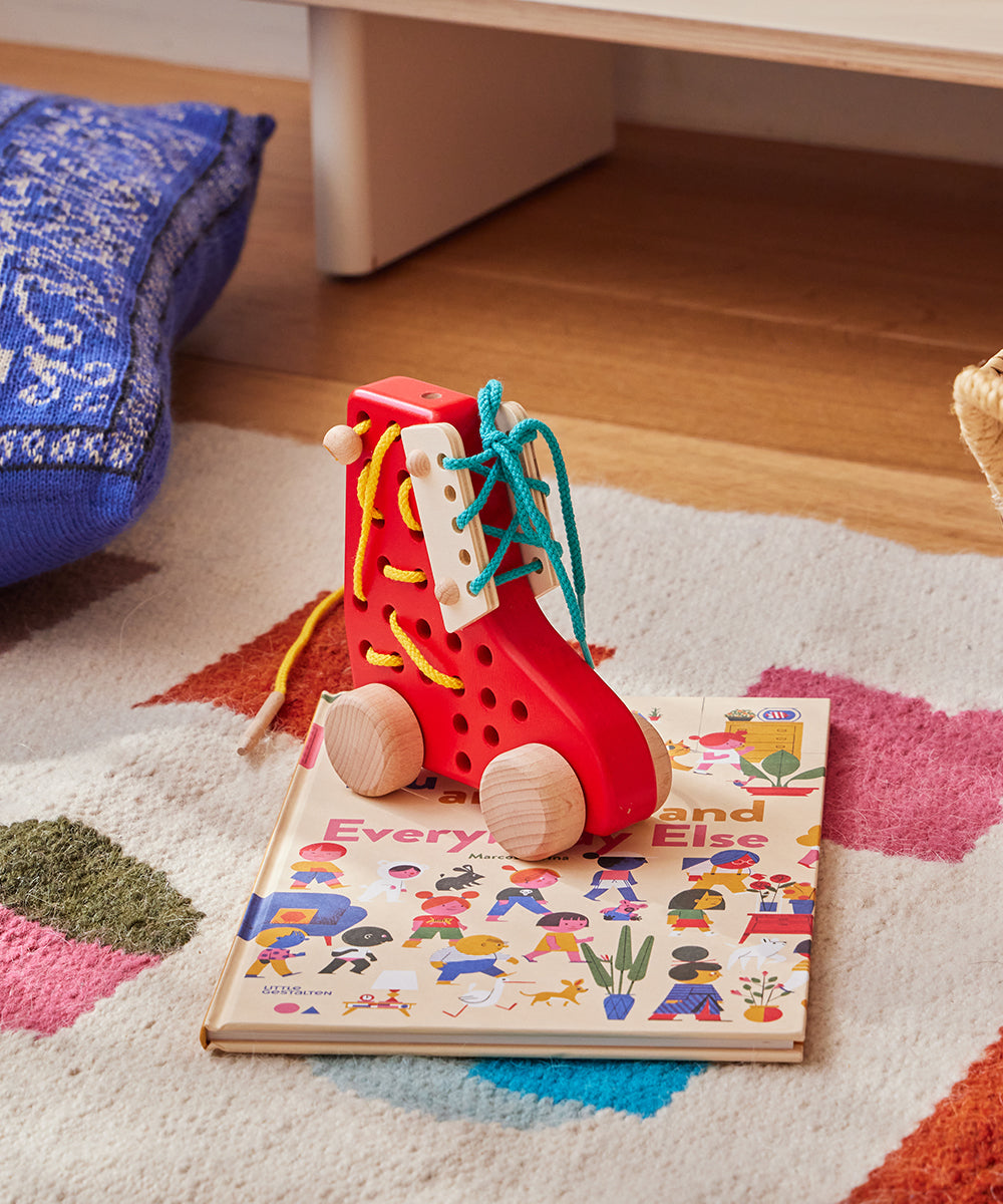 The Le Lacing Shoe, a red wooden toy with colored laces, is placed atop a children's book on a colorful rug.
