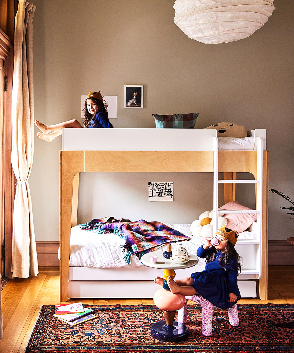 Two kids play happily in a cozy room with toys and books; one sits atop the Perch Twin Bunk Bed, the other on a flamingo chair, both smiling.