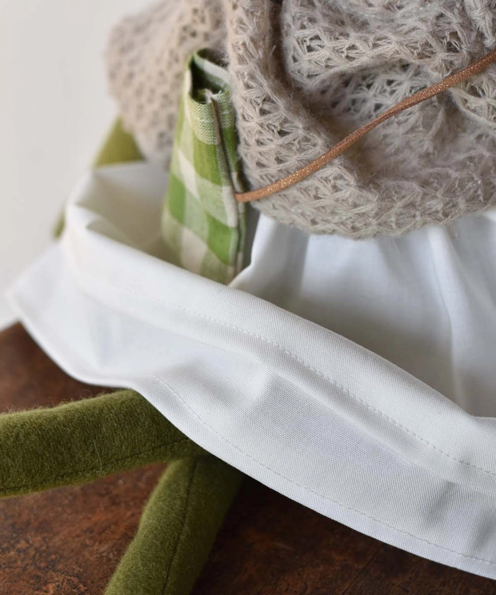 Close-up of Fern le Frog, a stuffed animal with a white skirt, green legs, and a green checkered pouch, sitting on wood.